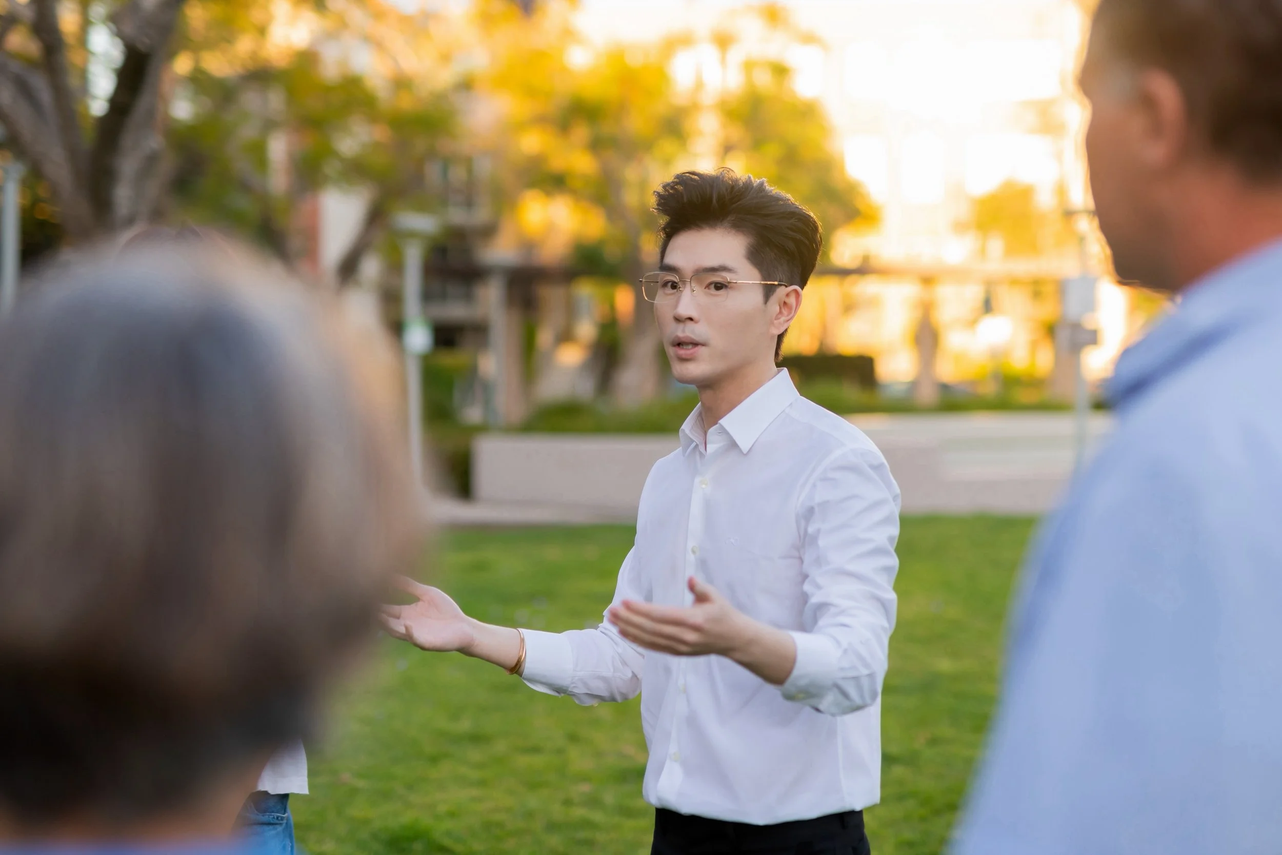 A young man wearing glasses and a white button-up shirt is talking to a group of people outdoors during sunset in a park, with trees and buildings in the background.