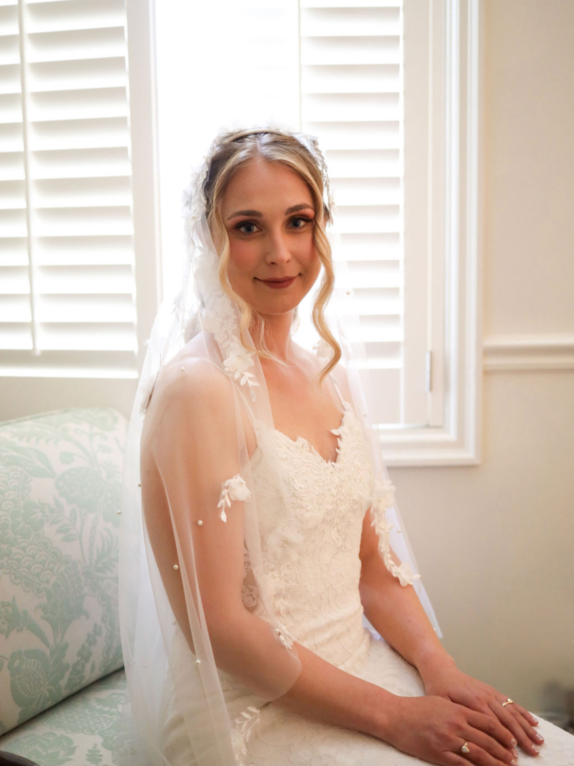 Bride with soft, natural glam makeup sitting by a window in her wedding dress, looking calm, radiant, and effortlessly beautiful before her ceremony