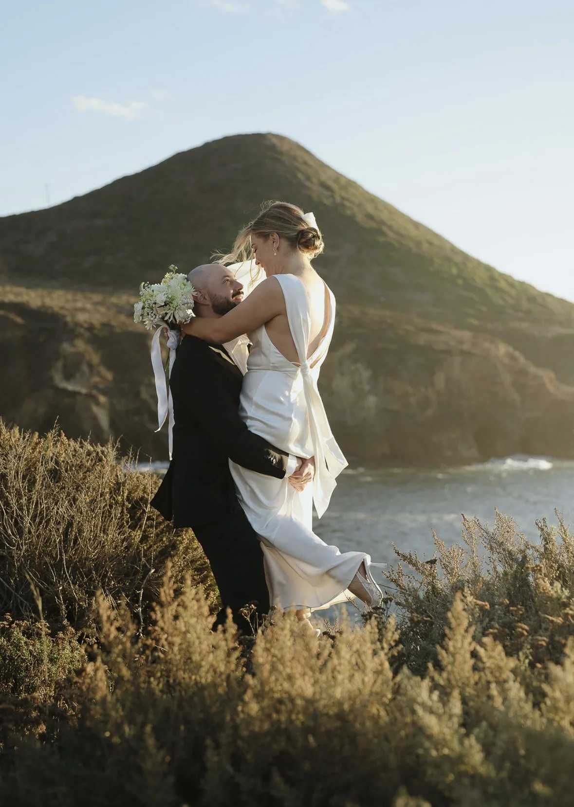 Bride with radiant, natural glam makeup lifted by her partner at sunset, glowing with confidence and joy in a breathtaking coastal setting