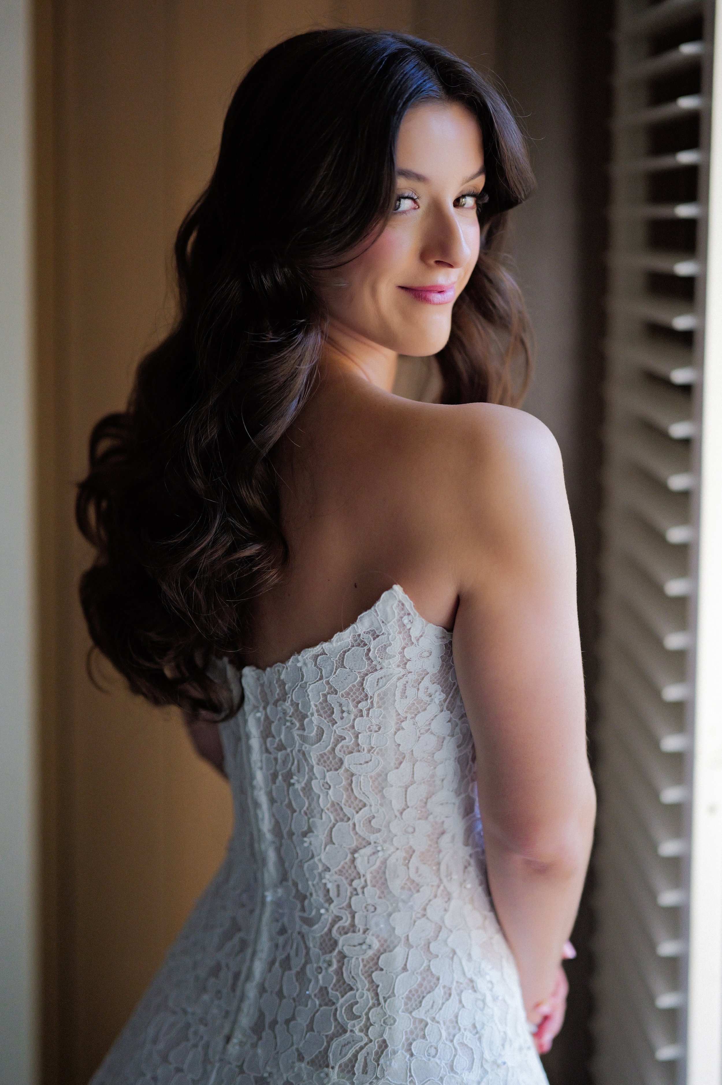 Bride with soft glam makeup and loose waves looking confident and radiant by the window before her wedding