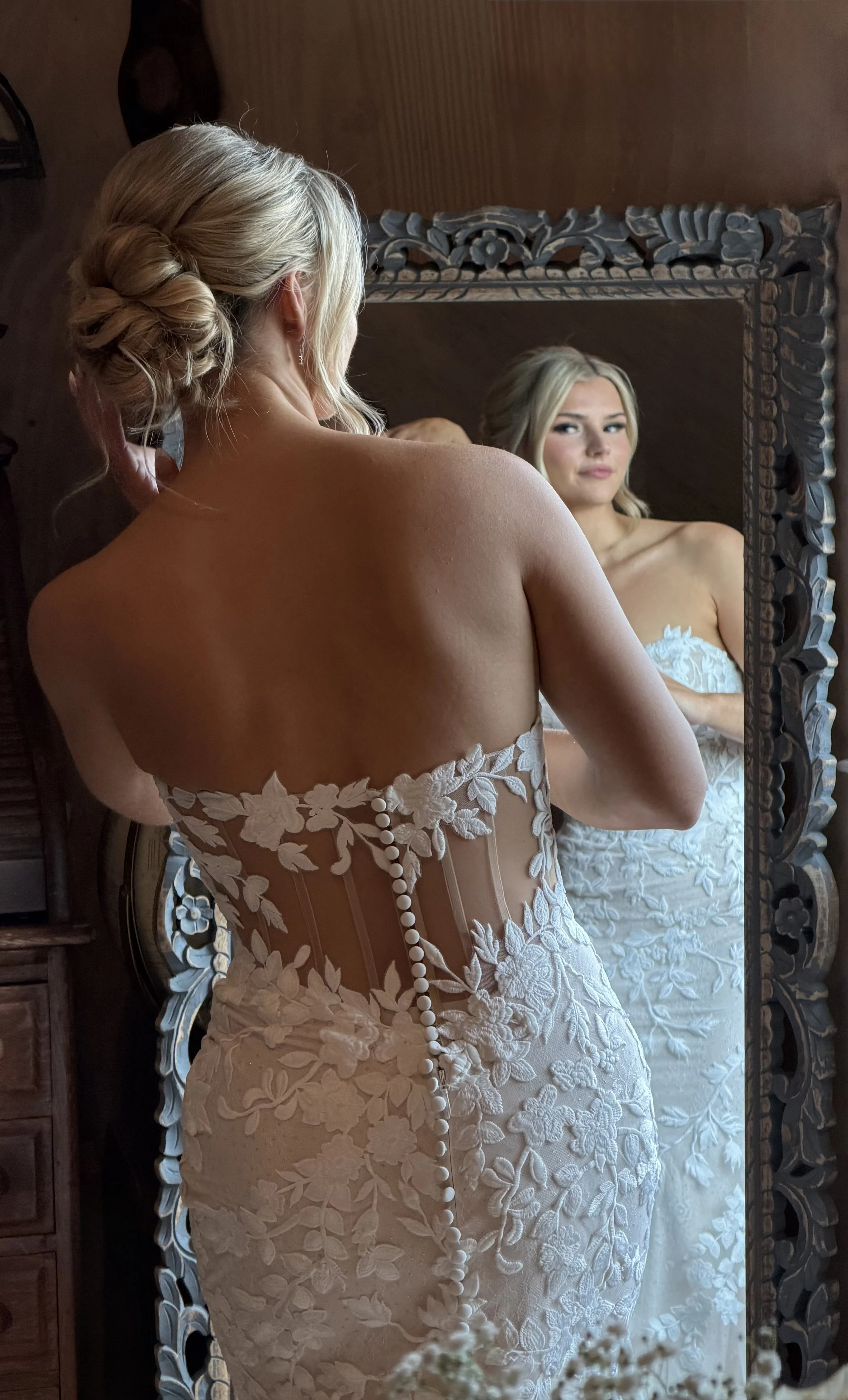 Bride with soft, timeless bridal glam and romantic updo adjusting her dress in the mirror, looking polished, confident, and wedding-day ready