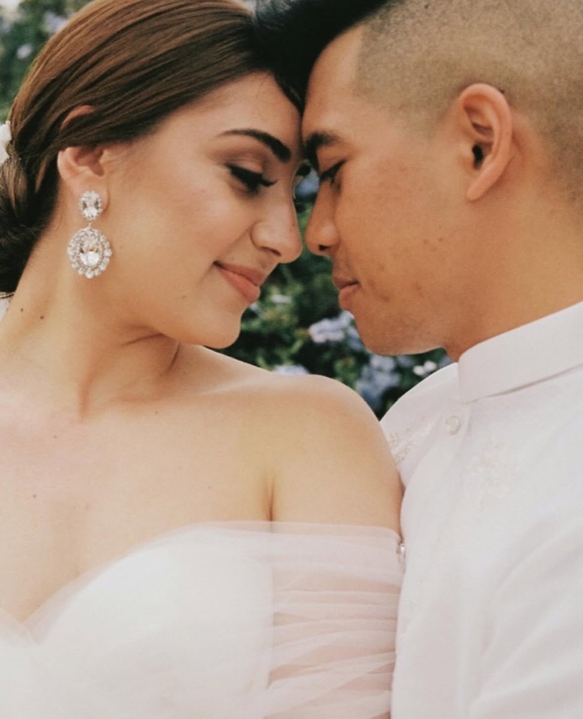 A close-up of a couple touching foreheads, smiling gently, outdoors with blurred greenery in the background in Baja CA MX.