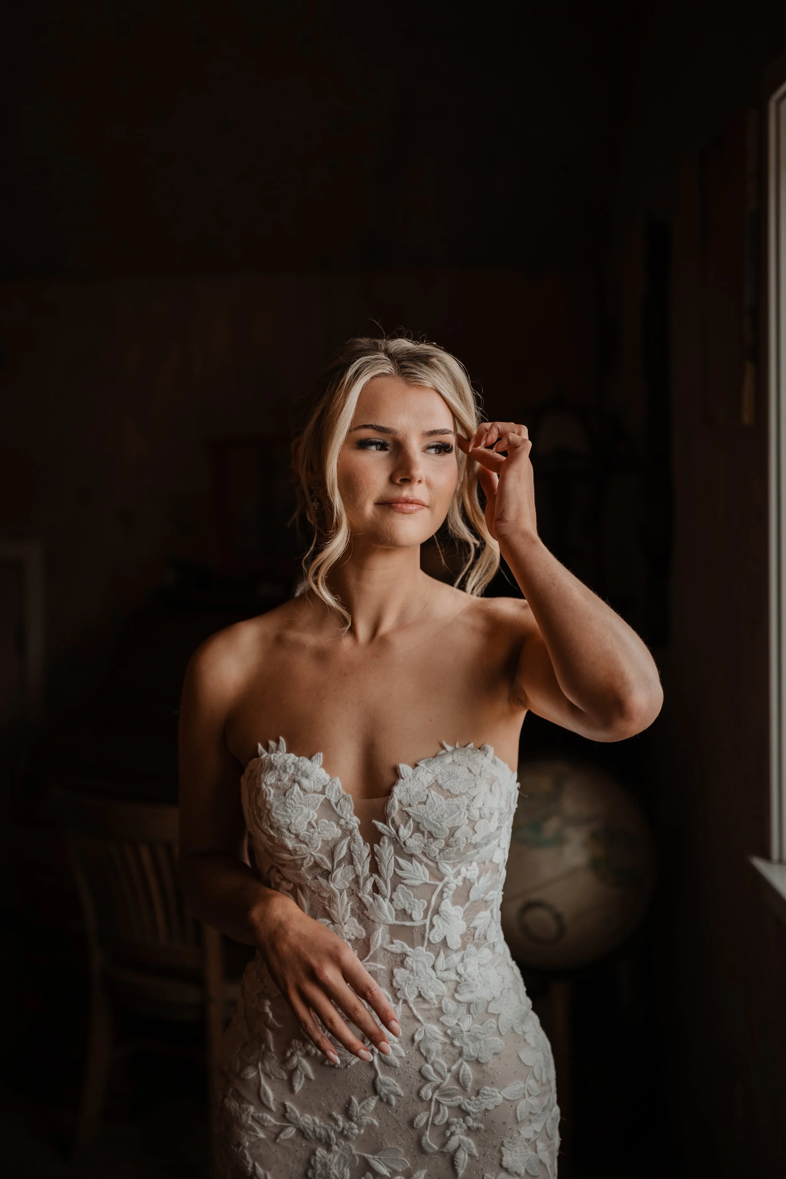 Bride with soft, timeless bridal glam adjusting her hair by the window, looking calm, confident, and radiant