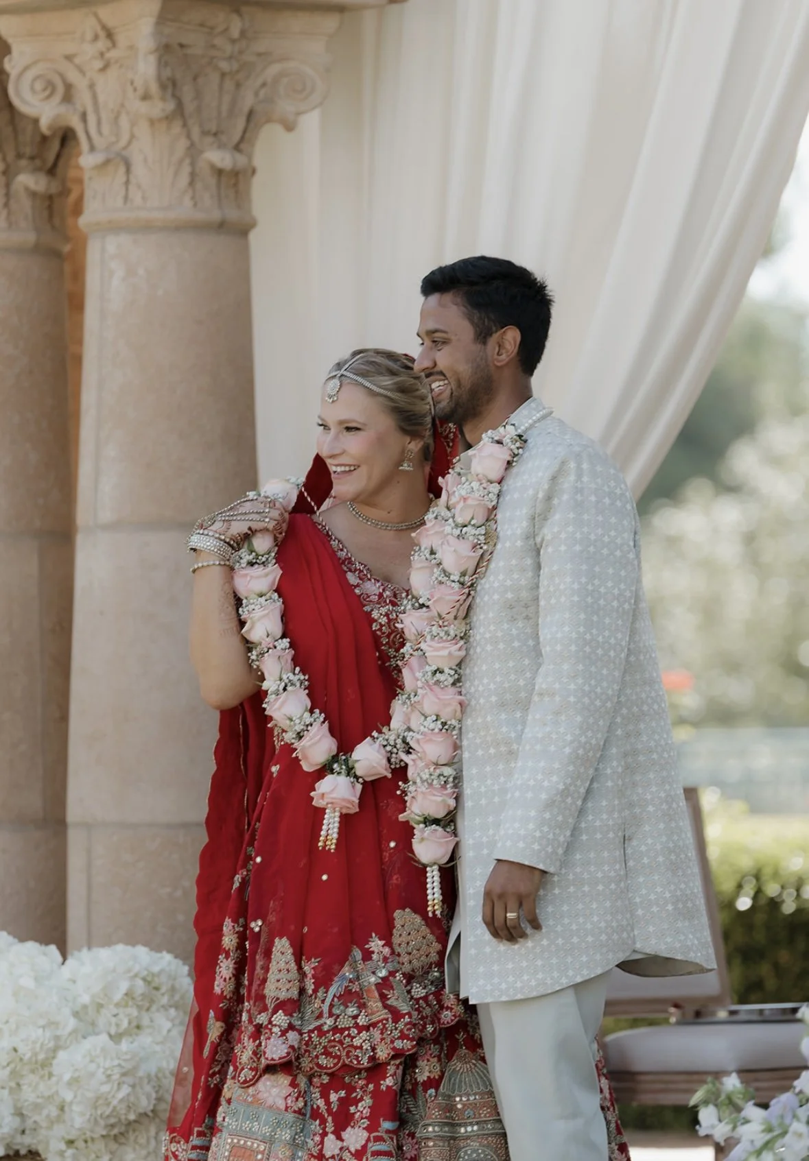 A bride and groom smiling and embracing at their wedding, with the bride wearing a red traditional South Asian dress and jewelry, and the groom in a white patterned sherwani, both adorned with floral garlands.