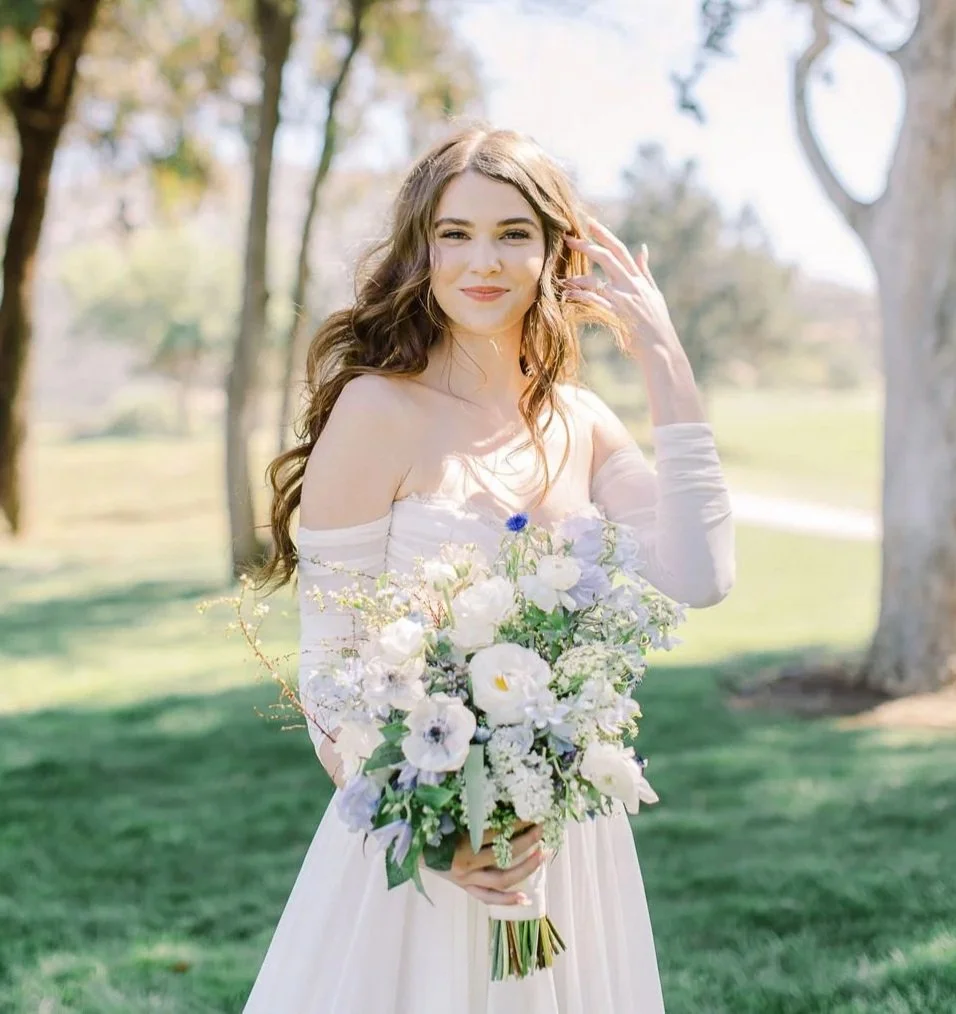 Bride with soft, natural glam makeup and romantic bridal hairstyle holding bouquet at an outdoor wedding