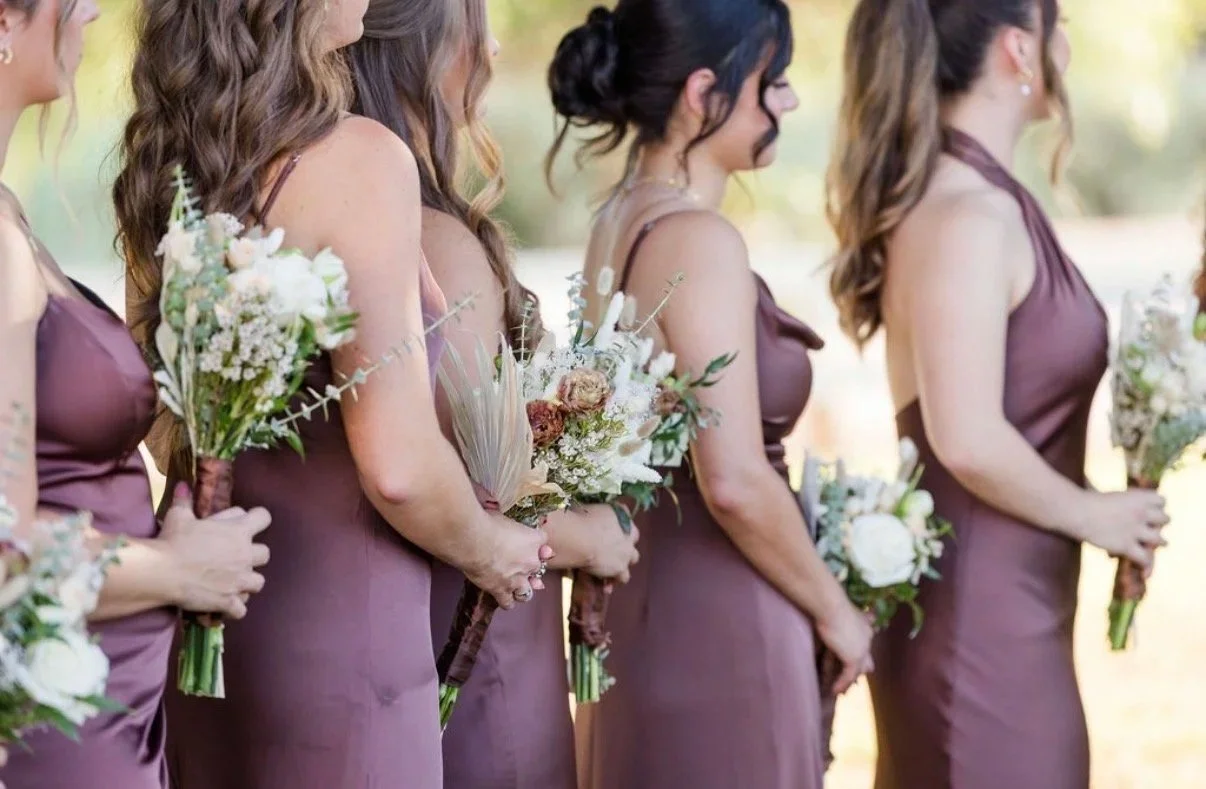 Bridesmaids with soft, cohesive hair and makeup looks holding bouquets at an outdoor wedding celebration