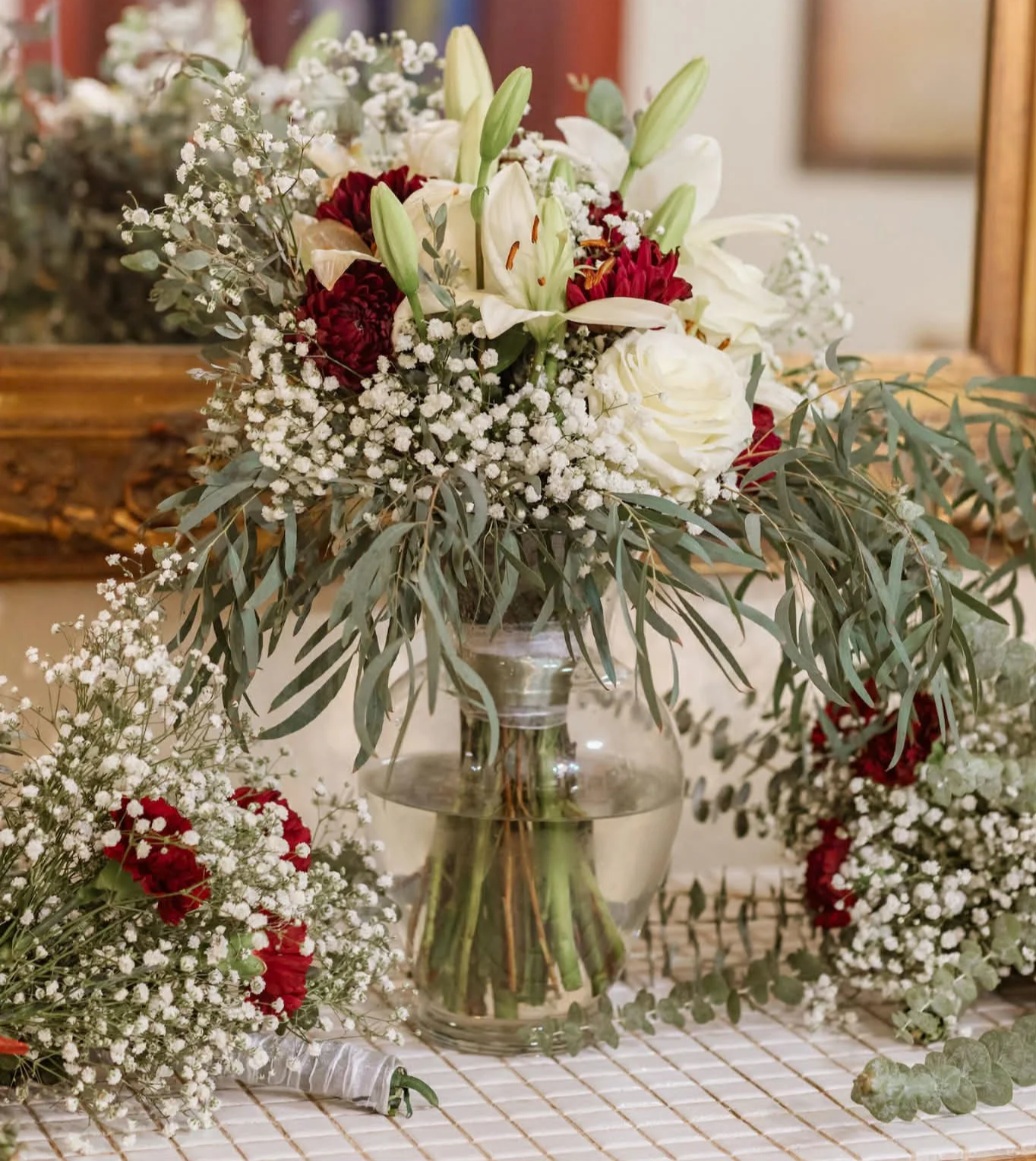 A floral arrangement in a glass vase with white lilies, red flowers, white roses, baby's breath, and greenery, placed on a table with additional floral decorations around it.