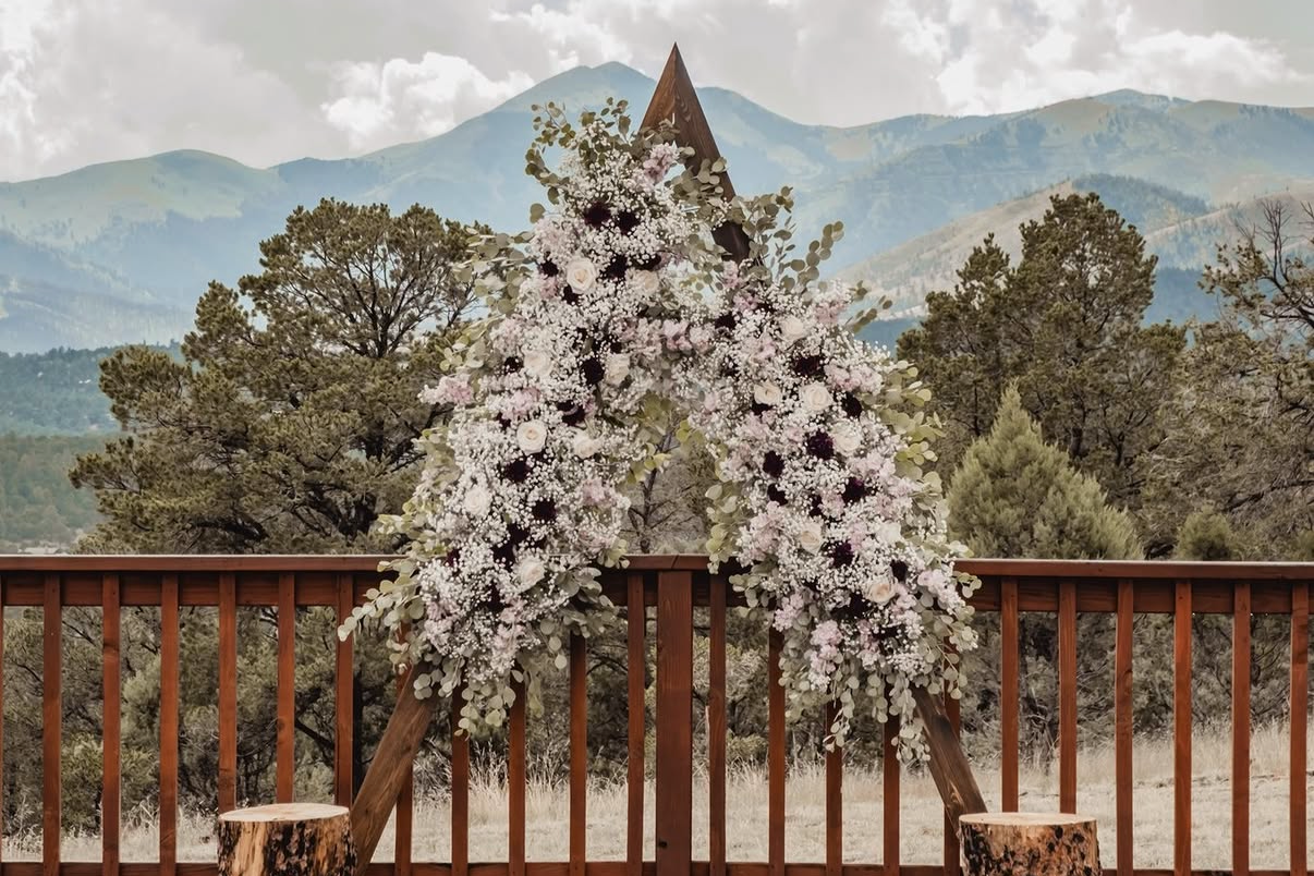 A floral wedding arch decorated with white, pink, and dark purple flowers, set on a wooden deck with mountain scenery in the background.
