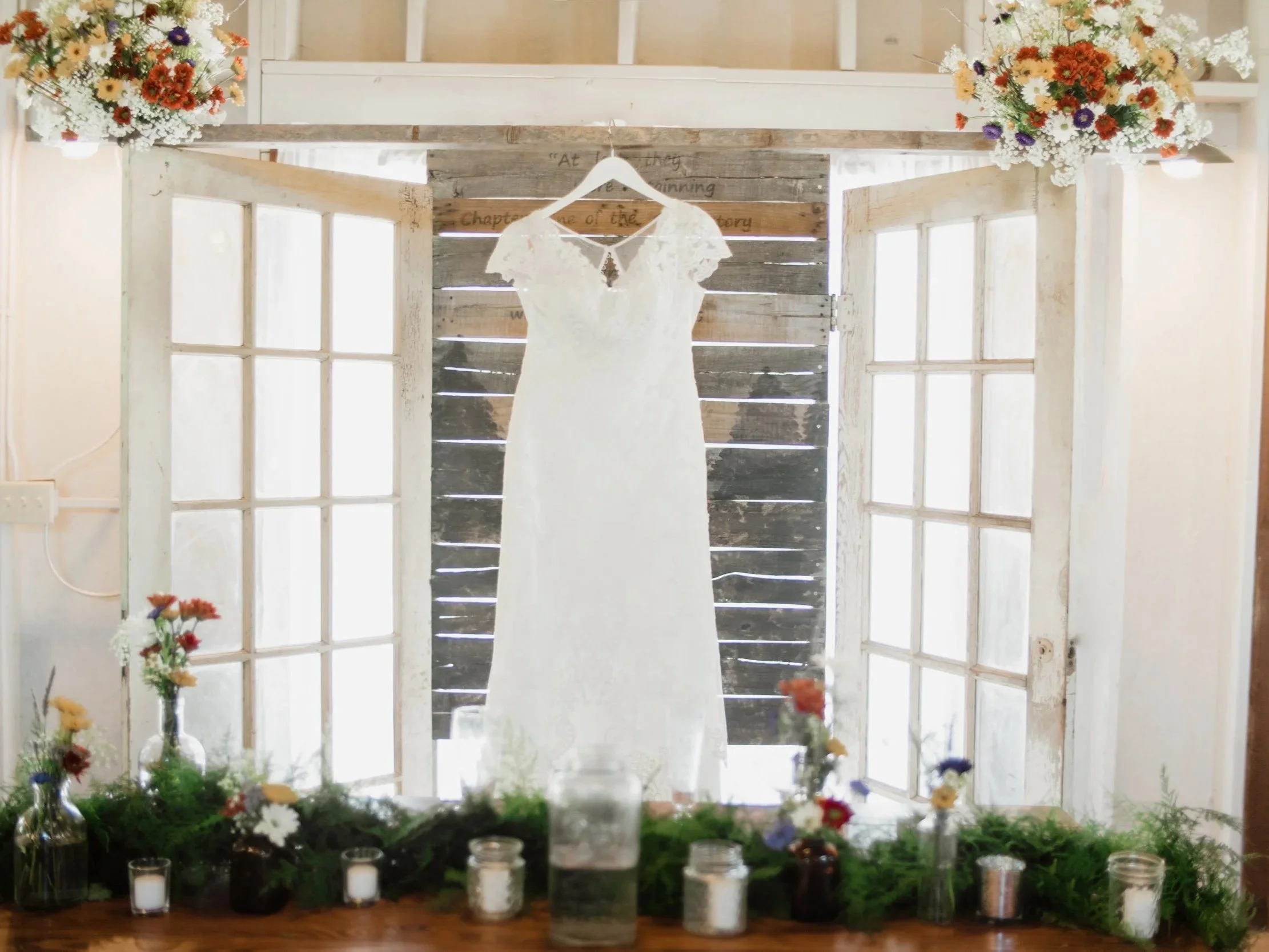 White wedding dress hanging on a wooden background inside a rustic wedding venue decorated with flowers on either side and candles along a table in the foreground.