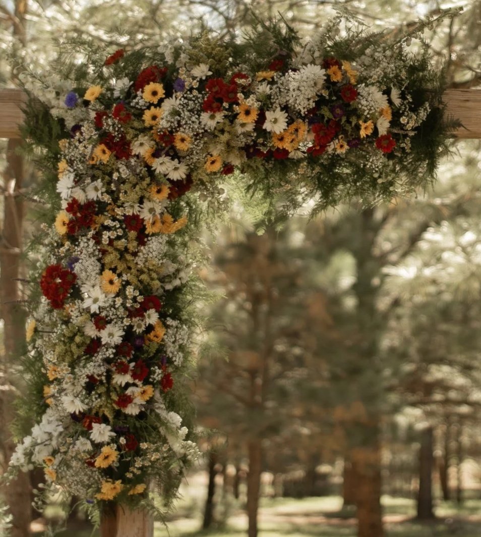 Colorful floral arrangement with various flowers and greenery, mounted on a wooden frame outdoors among trees.