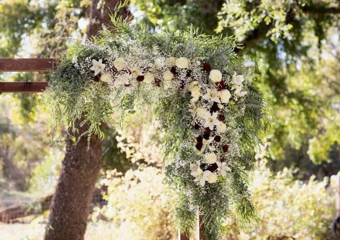 A floral wedding arch decorated with white and red flowers and green foliage, set outdoors with trees in the background.