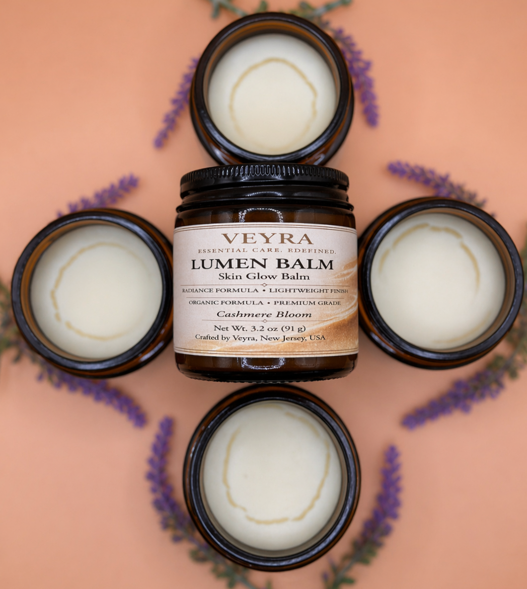 Four brown jars with white balm inside, one jar opens showing the balm, surrounded by purple lavender flowers on a pink background. The center jar's label reads 'Veyra Lumen Balm, Skin Glow Balm, Cashmere Bloom, 3.2 oz, crafted in New Jersey, USA.'