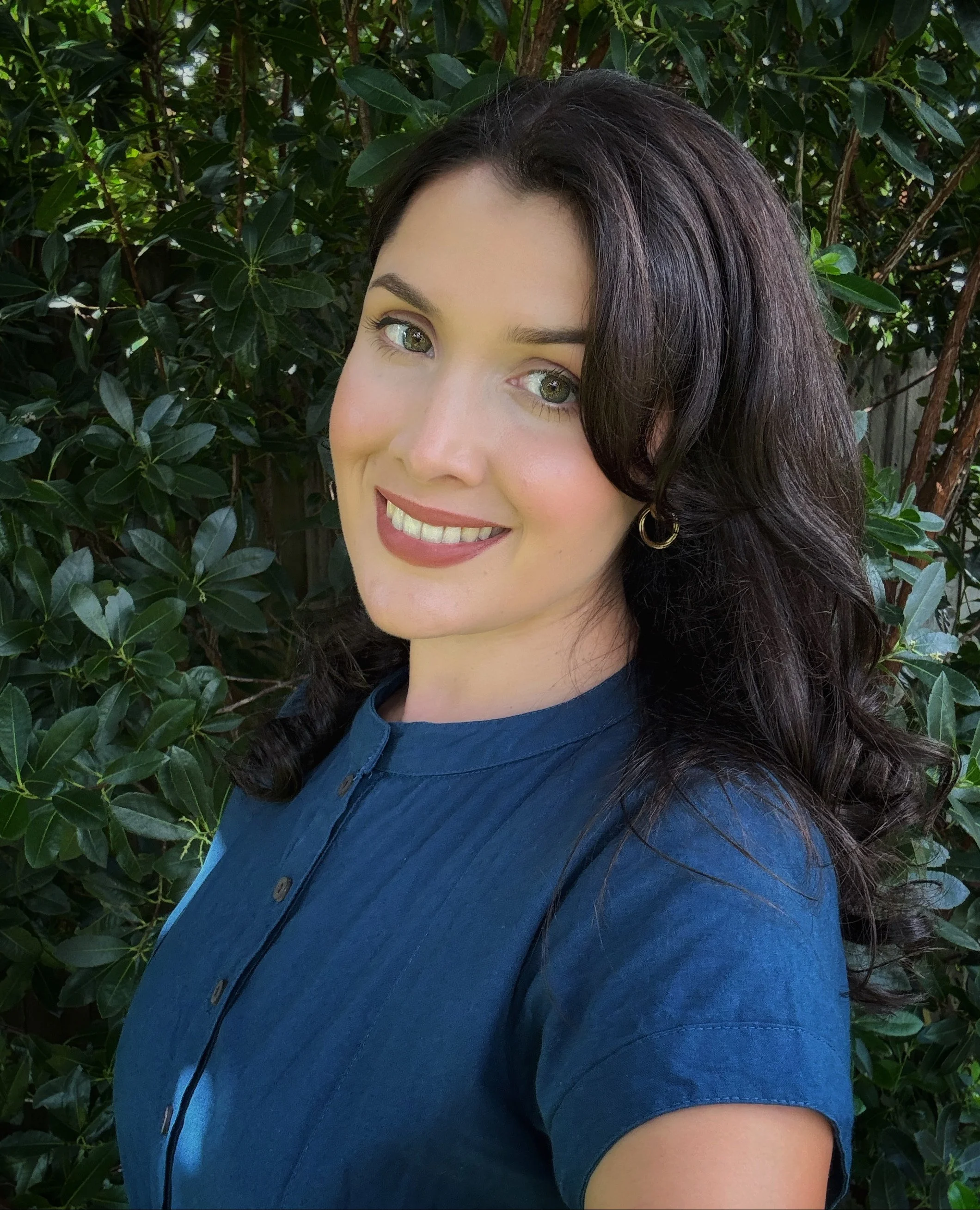 Close-up of a woman with long dark hair and light skin, smiling, outdoors near green leafy bushes.