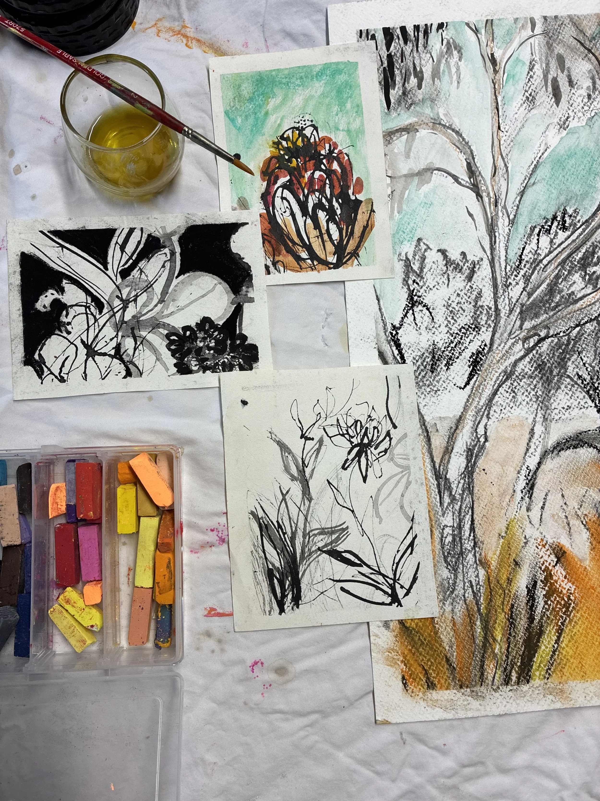 Displays various paintings and drawings of flowers and nature scenes created with watercolor and ink, along with pastel chalks in a plastic container, on a white table covered with paint splashes.