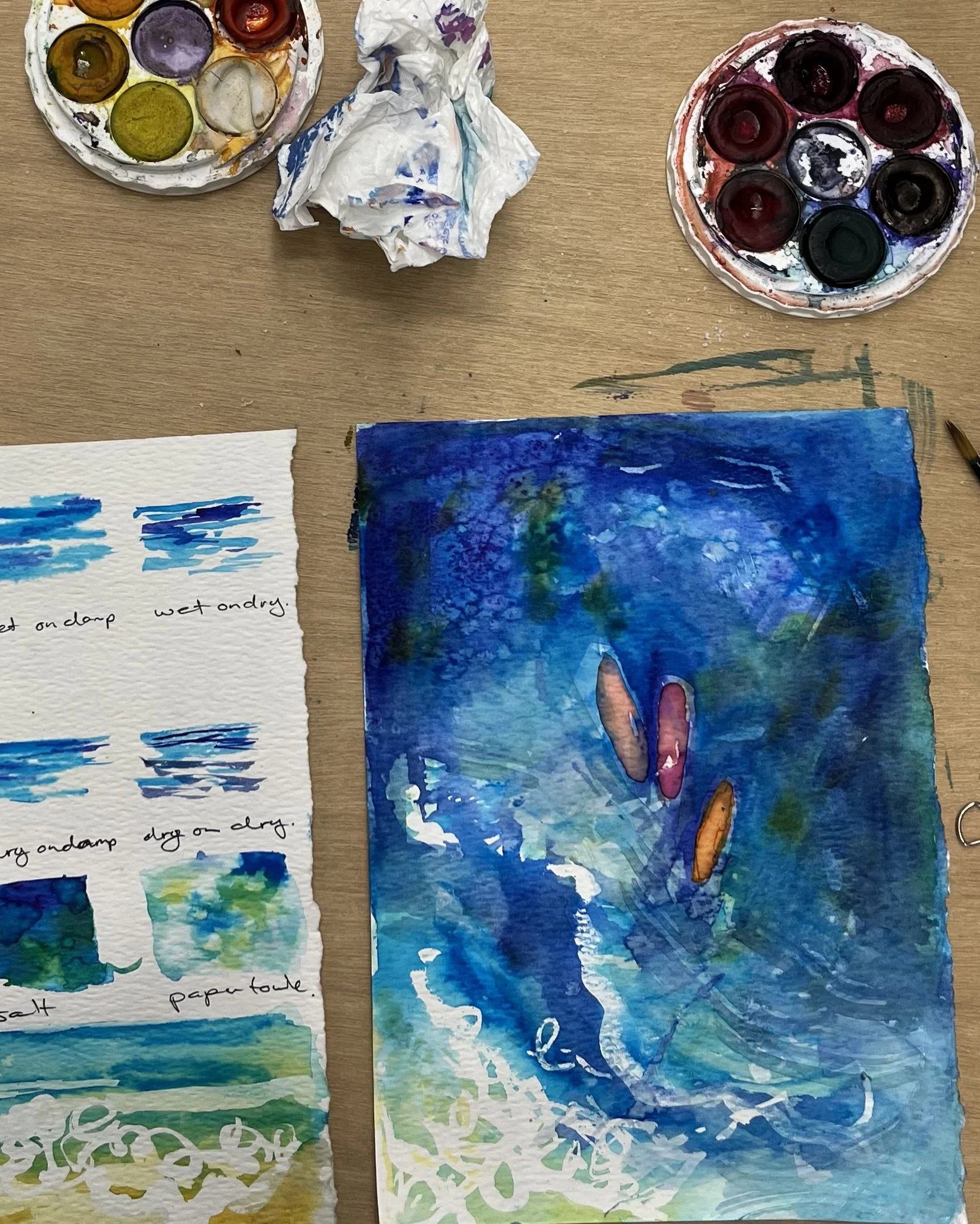 Watercolor paintings of water scenes, featuring blue and green hues with some aquatic elements, on textured watercolor paper. There are also circular watercolor palettes containing various colors and some crumpled tissue paper on the wooden surface.