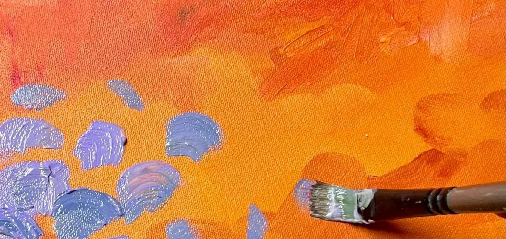 Close-up of an abstract painting in progress, featuring orange background with purple and light pink brushstrokes and a flat paintbrush applying white paint.