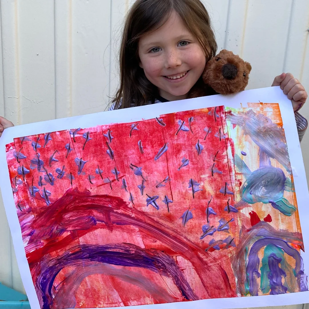 A young girl with brown hair and freckles smiling while holding a teddy bear and a colorful painting.