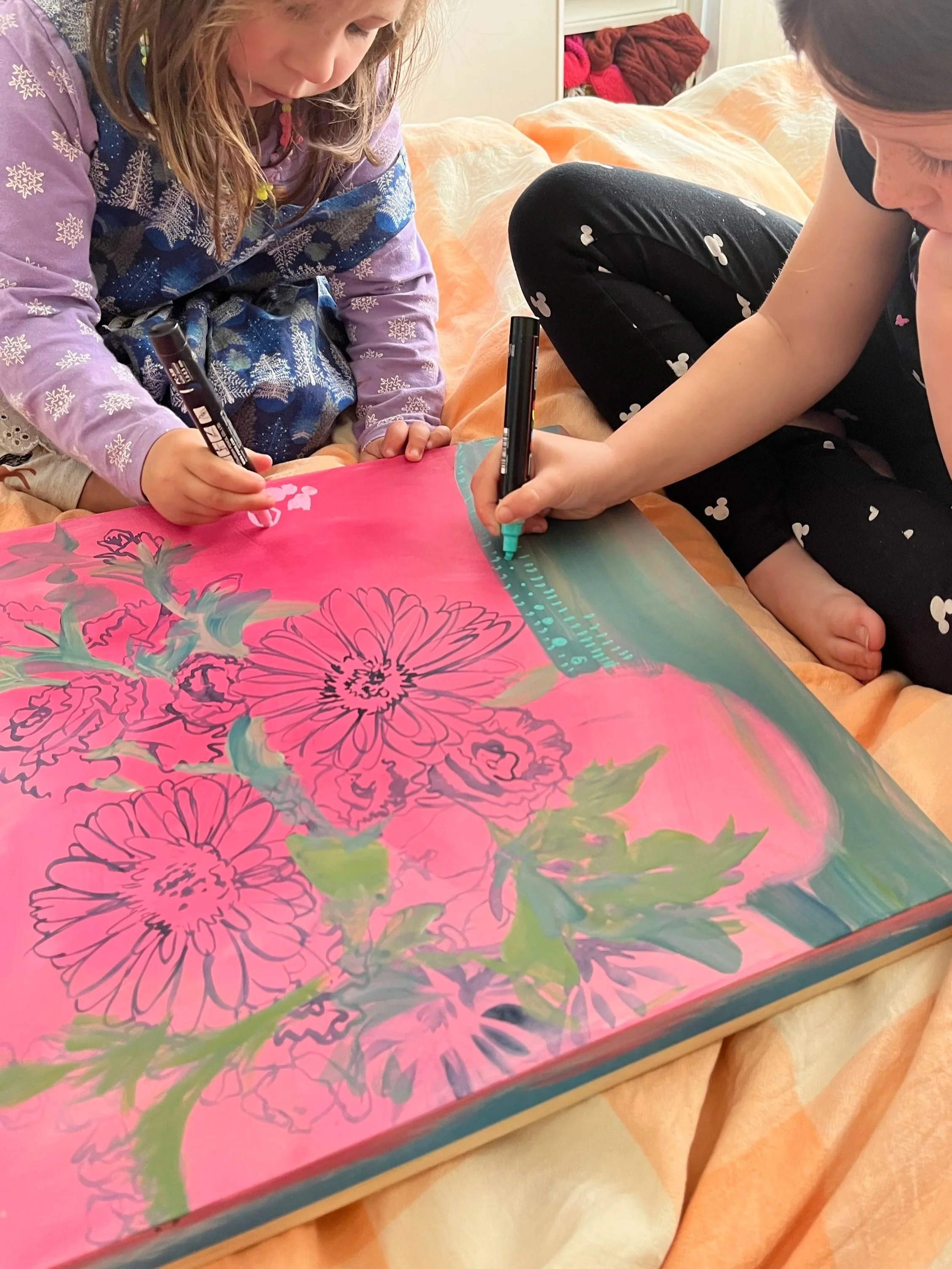 Two children are sitting on a bed while drawing and coloring on a large piece of pink paper with floral designs, using black markers.