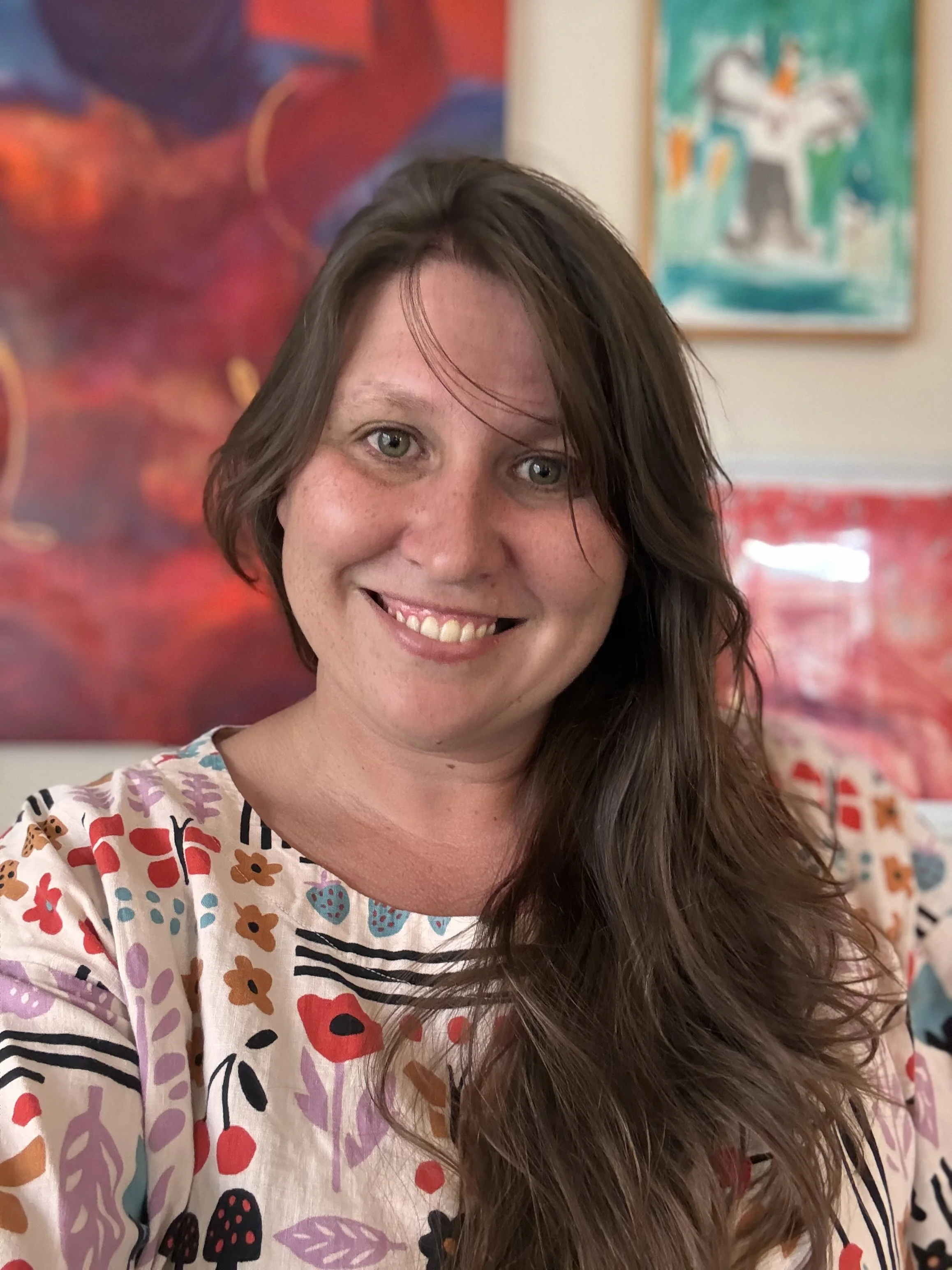 A woman with long, wavy brown hair smiling, wearing a colorful short-sleeve shirt with floral and fruit patterns, with artwork and colorful paintings in the background.