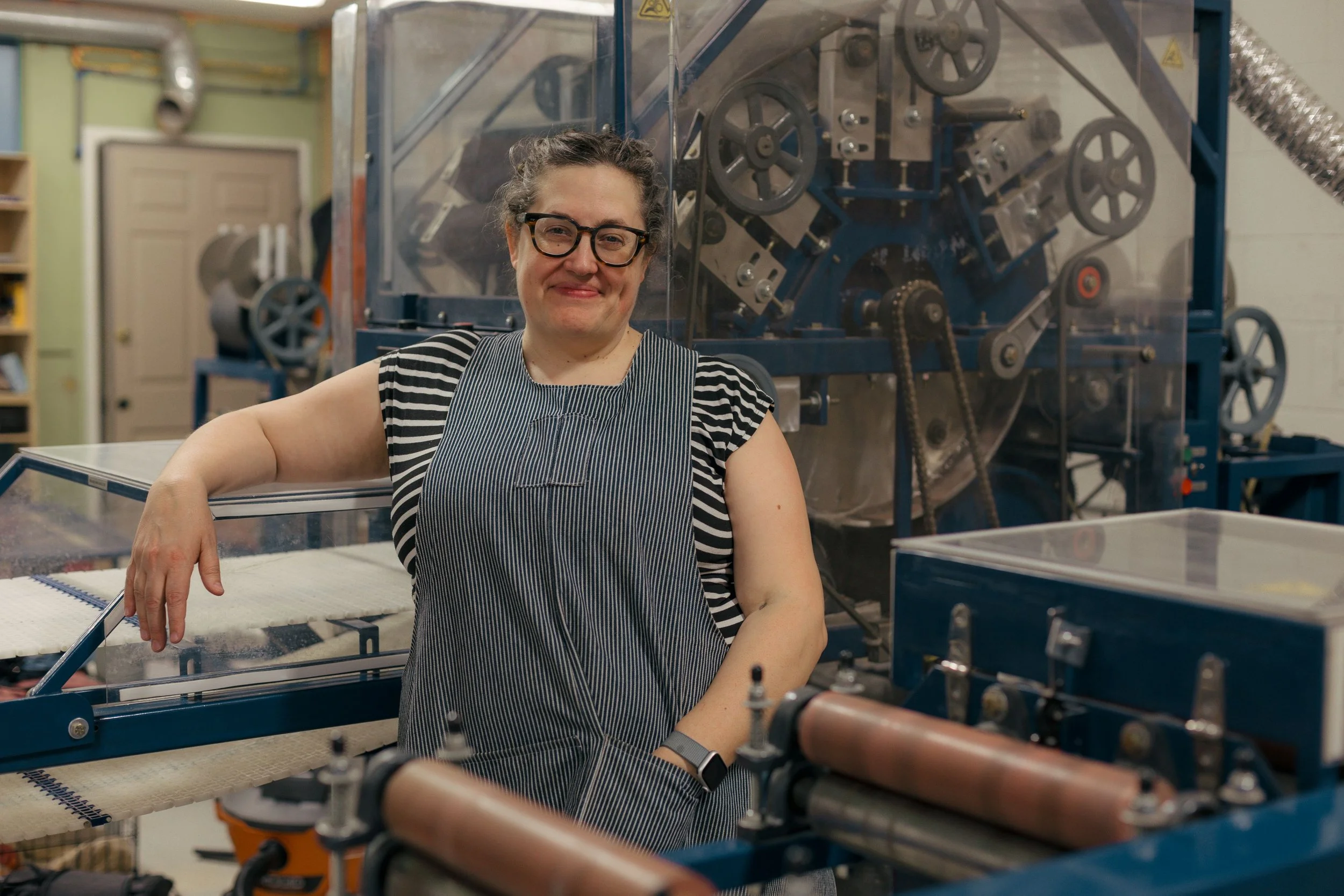 A woman with glasses and a striped shirt smiling while working with machinery in a factory or workshop setting.