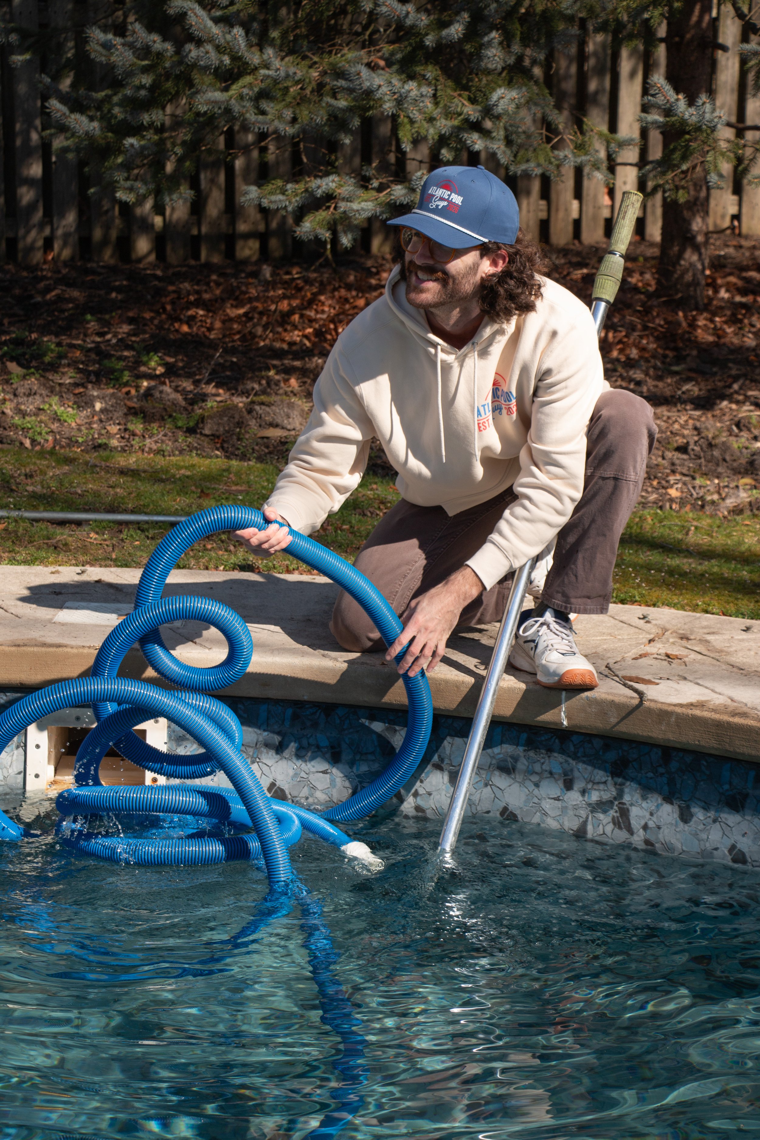 A man wearing a beige hoodie with a colorful Atlantic Pool Guys logo on the back, on his knees by a swimming pool, holding a blue pool vacuum hose, with a pool pole in his other hand, surrounded by trees and a wooden fence.