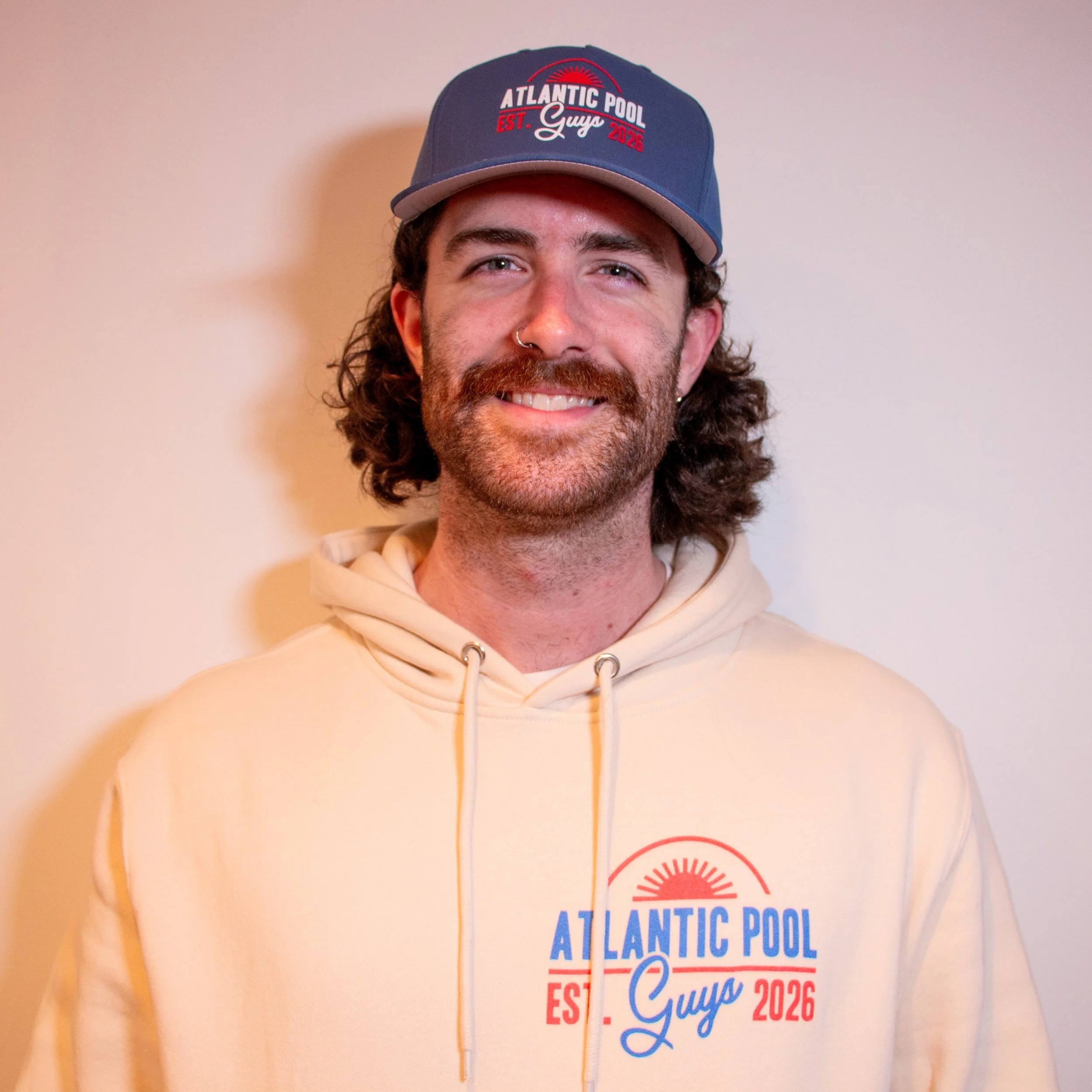 A man with curly hair and a beard wearing a blue and gray baseball cap and a beige hoodie. The cap has the text 'Atlantic Pool Guys Est. 2026' and the hoodie has the same logo.