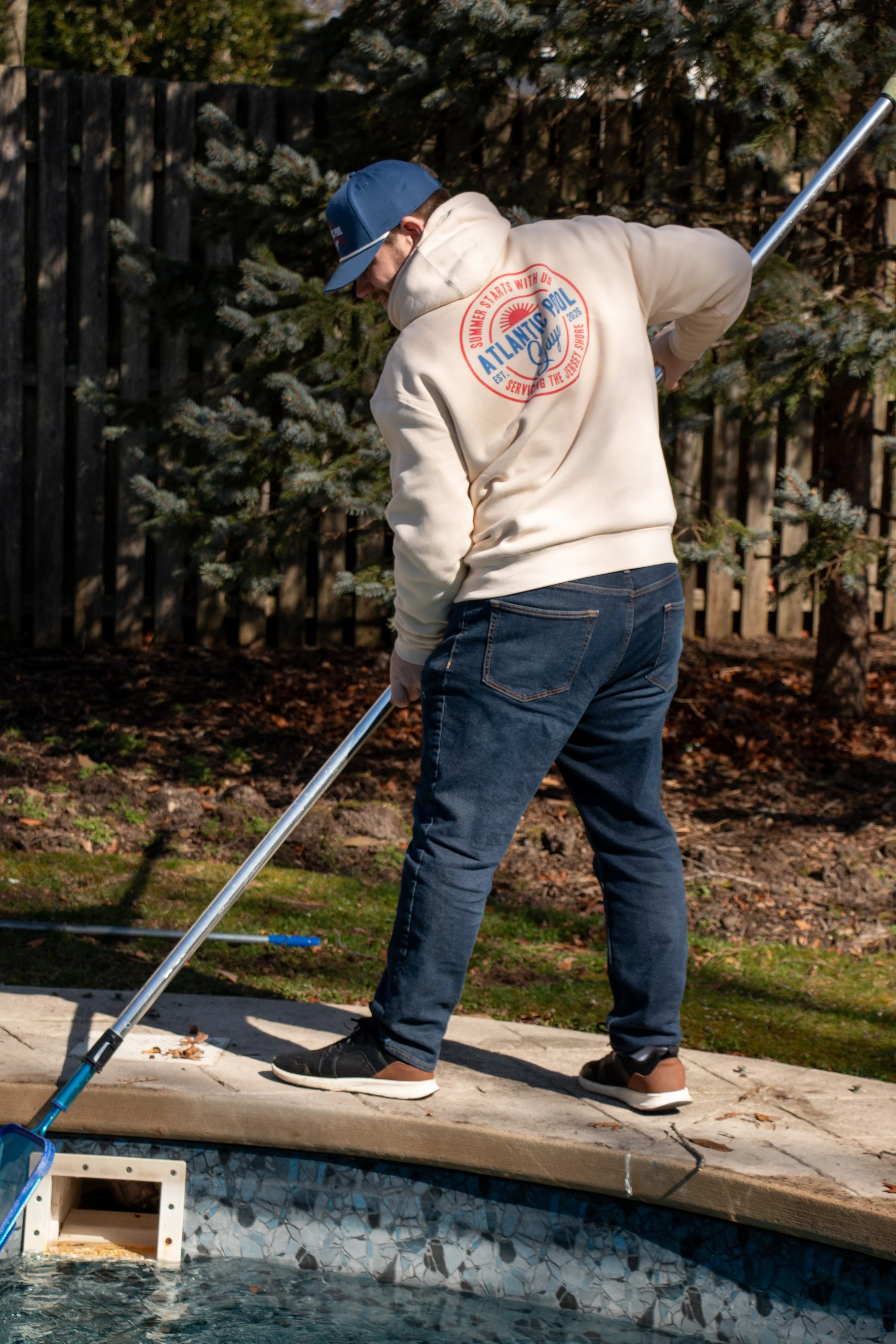 A person wearing a beige hoodie with a colorful Atlantic Pool Guys logo on the back, a blue baseball cap, jeans, and sneakers cleaning a backyard swimming pool using a long pole with a net.
