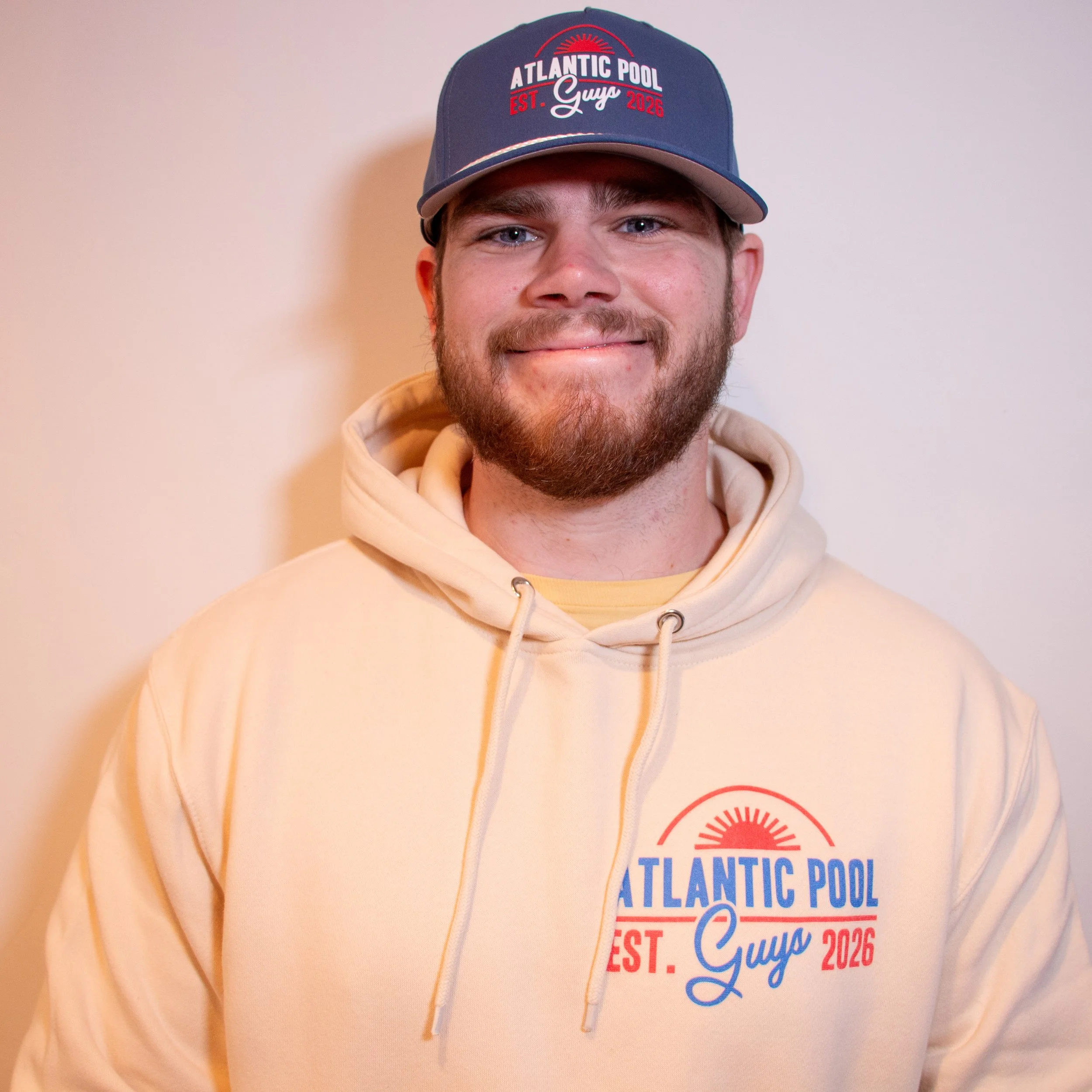 A young man wearing a navy blue baseball cap with red and white text and a beige hoodie with a similar logo. He is smiling and has a beard, standing against a plain light-colored wall.