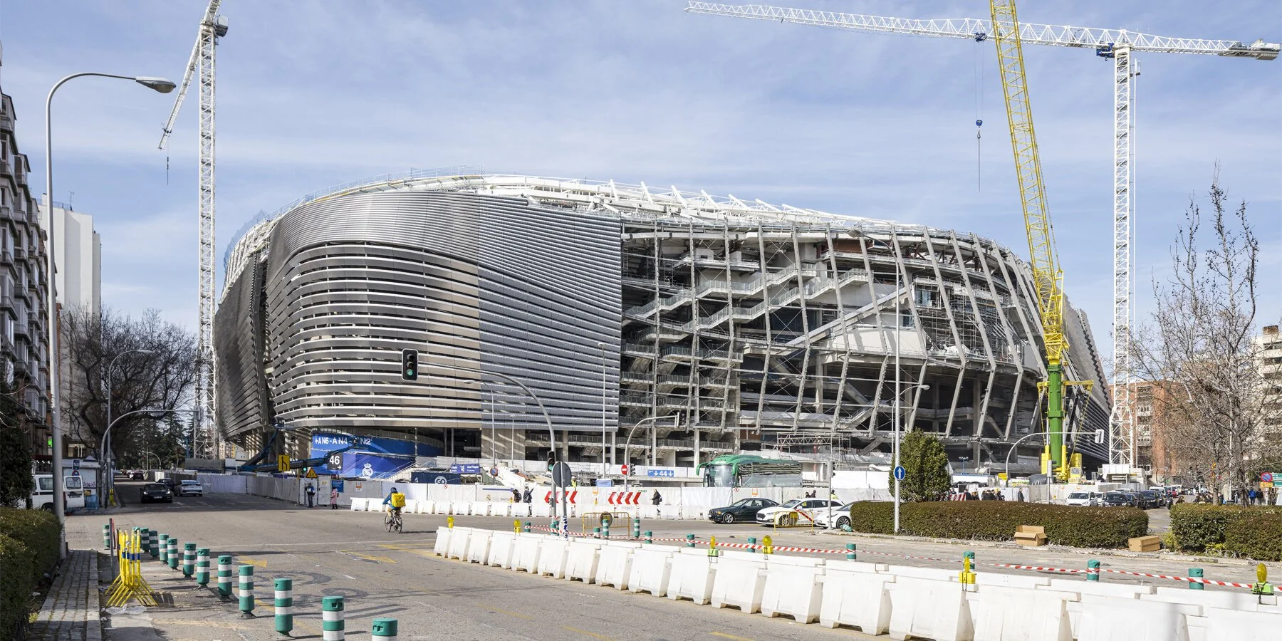 Under construction stadium with large yellow tower crane, modern curved architectural design, and scaffolding, located beside city streets with cars and pedestrians.