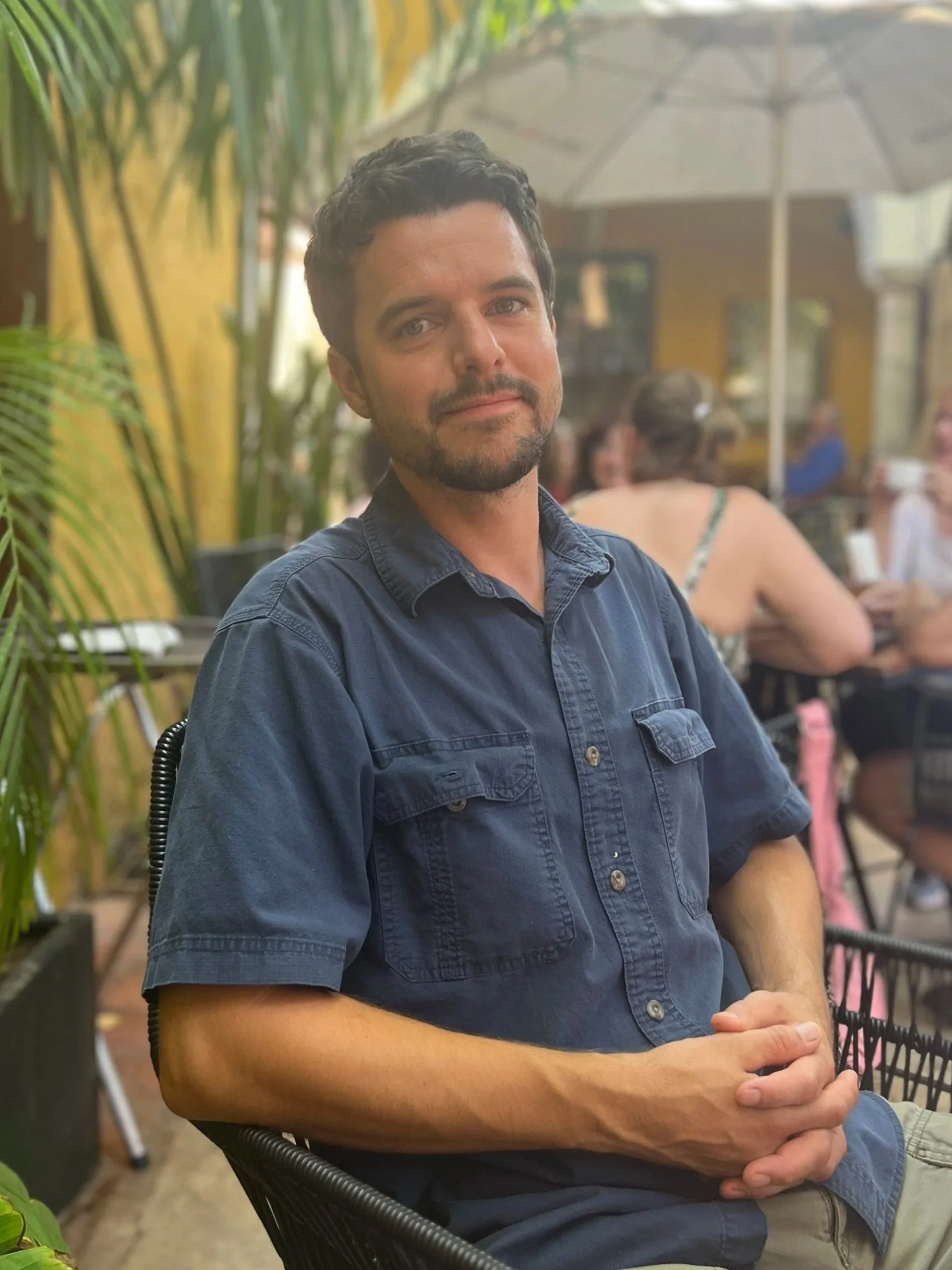 A man with dark hair and a beard wearing a navy shirt sitting at an outdoor cafe with greenery and other people in the background.