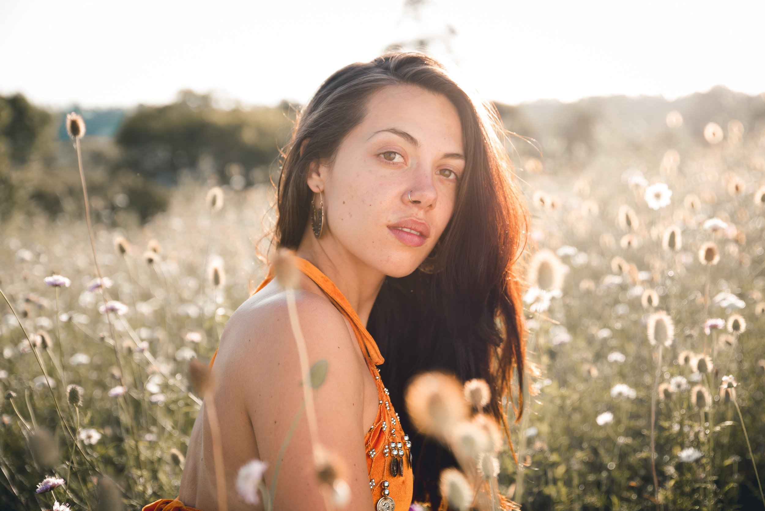 A young woman with long dark hair, wearing an orange dress with embellishments, sitting in a field of wildflowers at sunset.