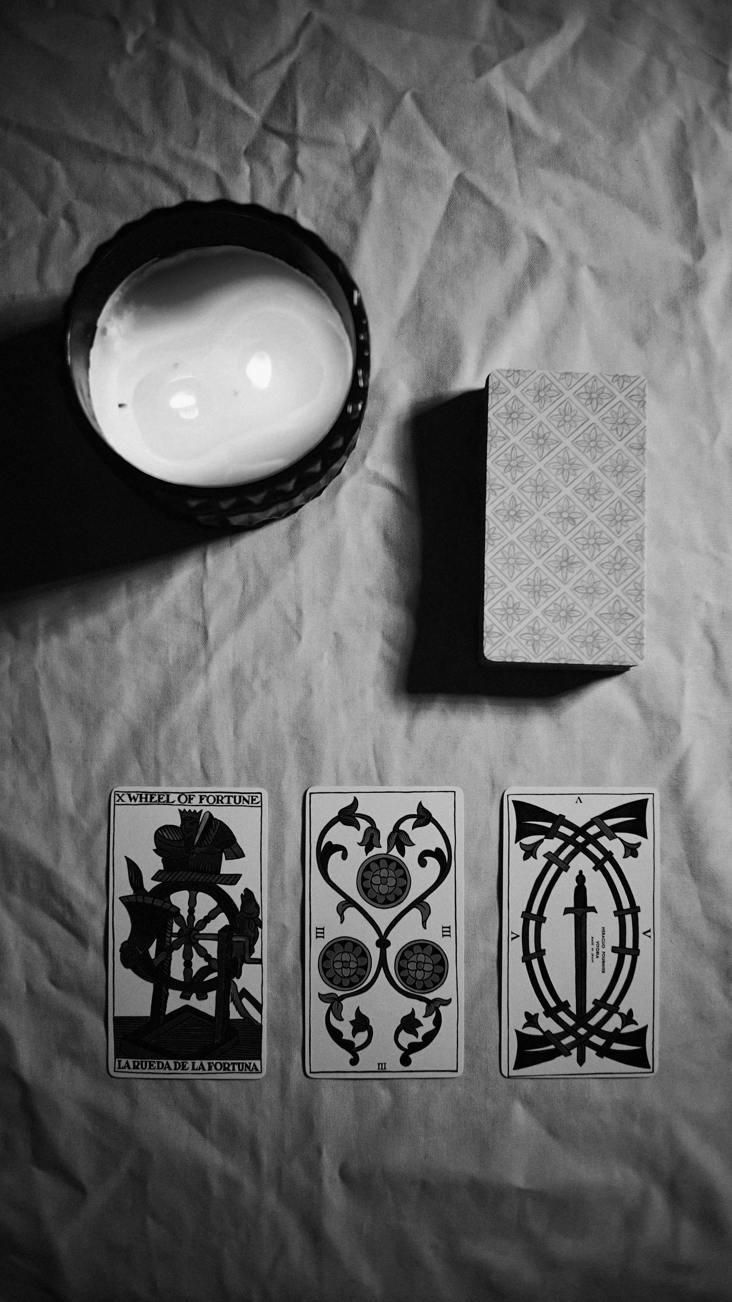 A lit candle in a textured glass holder, a deck of cards with a patterned back, and three tarot cards laid out on a wrinkled cloth surface.