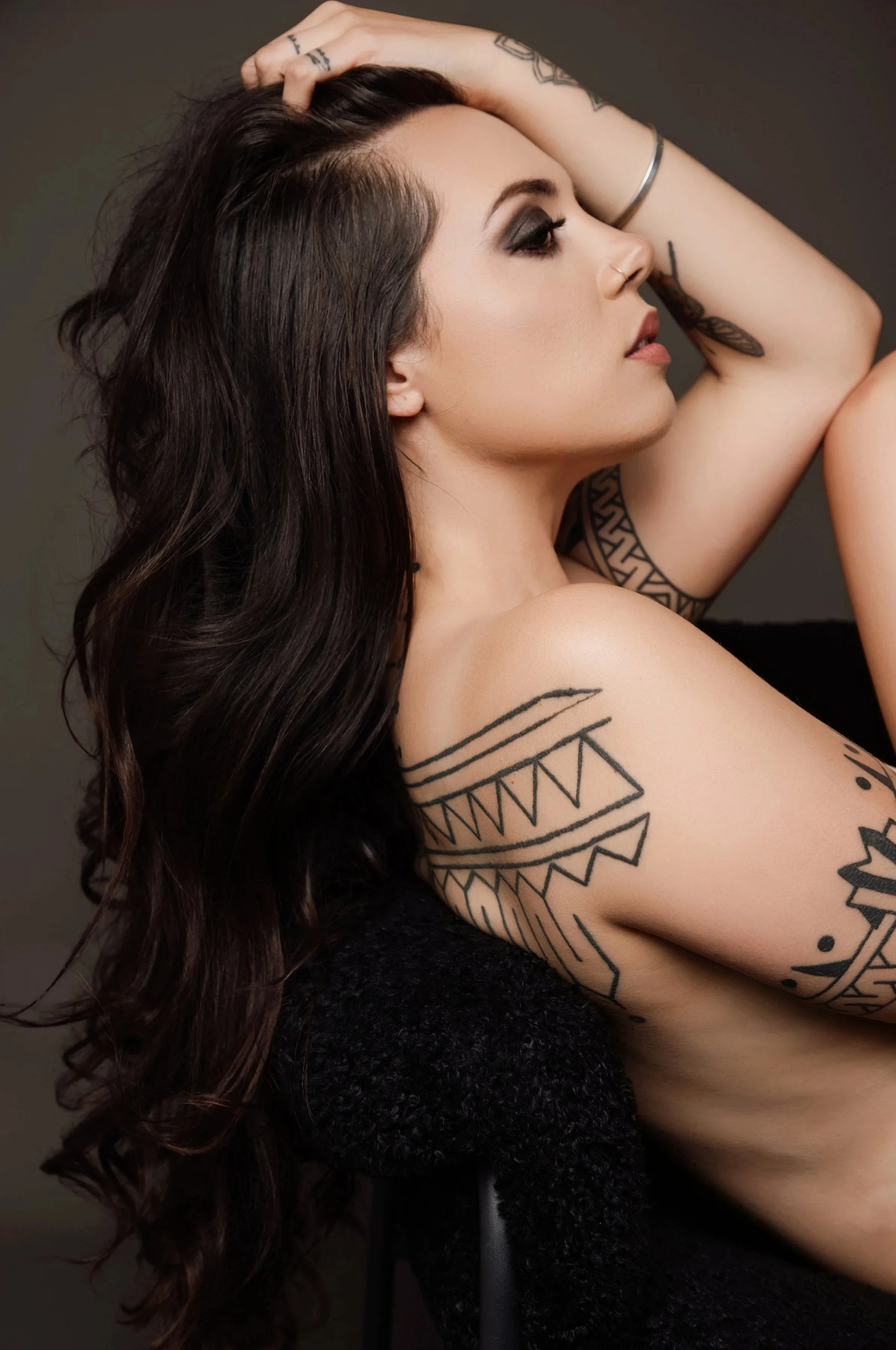 Side profile of a woman with long dark wavy hair, tattoos on her arm and shoulder, and dark makeup, sitting against a dark background.