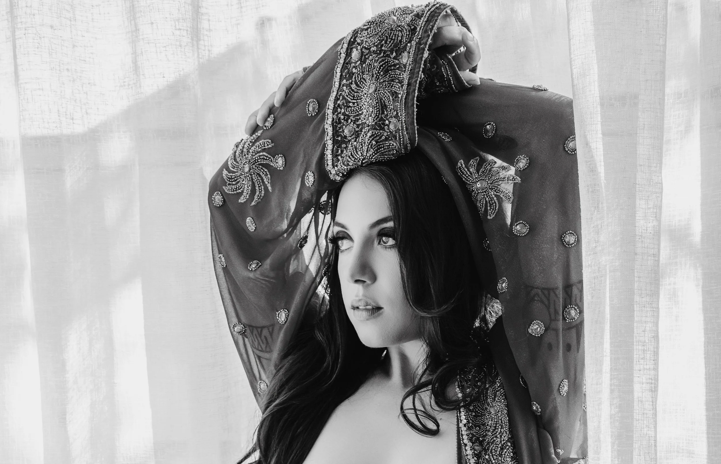 A woman with long dark hair looks to the side as she is lifting a sheer, embroidered fabric over her head, standing in front of a window with light filtering through curtains.