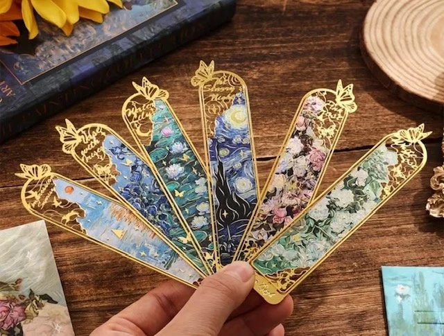 Hand holding six decorative bookmarks with floral and sky themes, featuring gold borders and intricate designs, on a wooden table.