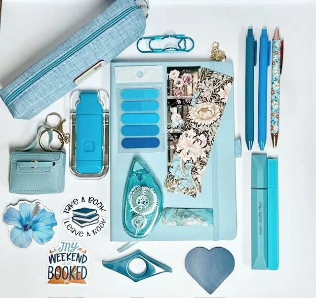 Flat lay of blue stationery and accessories, including pens, envelopes, a stapler, a pin, a notebook, stickers, and decorative items on a white background.