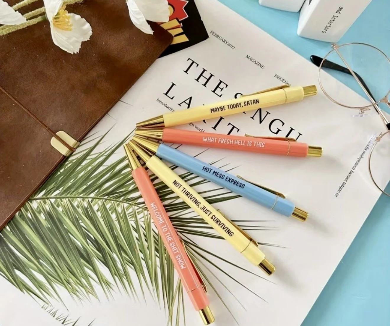 Six colorful pens with humorous labels on a desk, next to a magazine, a pair of round sunglasses, a brown notebook, and some papers with tropical leaf designs.