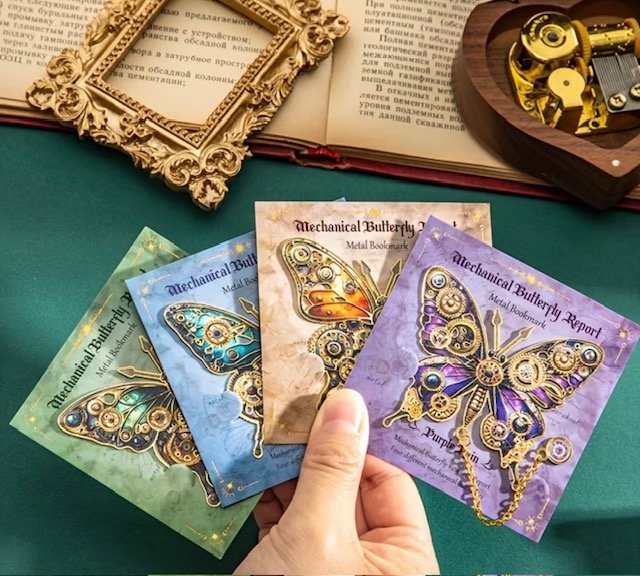 A hand holding four colorful mechanical butterfly metal bookmark cards with intricate gear designs, labeled as "Mechanical Butterfly" and "Mechanical Butterfly Report," on a green surface. In the background, an ornate empty picture frame, an open boo