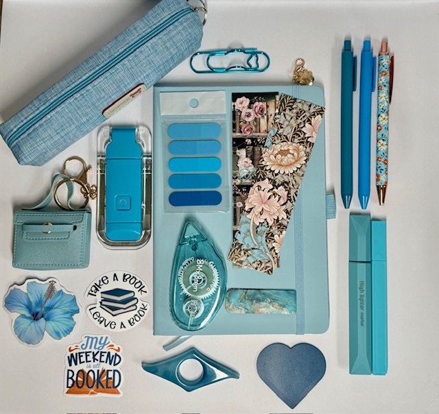 Collection of blue stationery and accessories including pens, a pencil case, a wallet, sticky notes, decorative tapes, a paper cutter, a heart-shaped item, a flower, and motivational stickers arranged on a white background.
