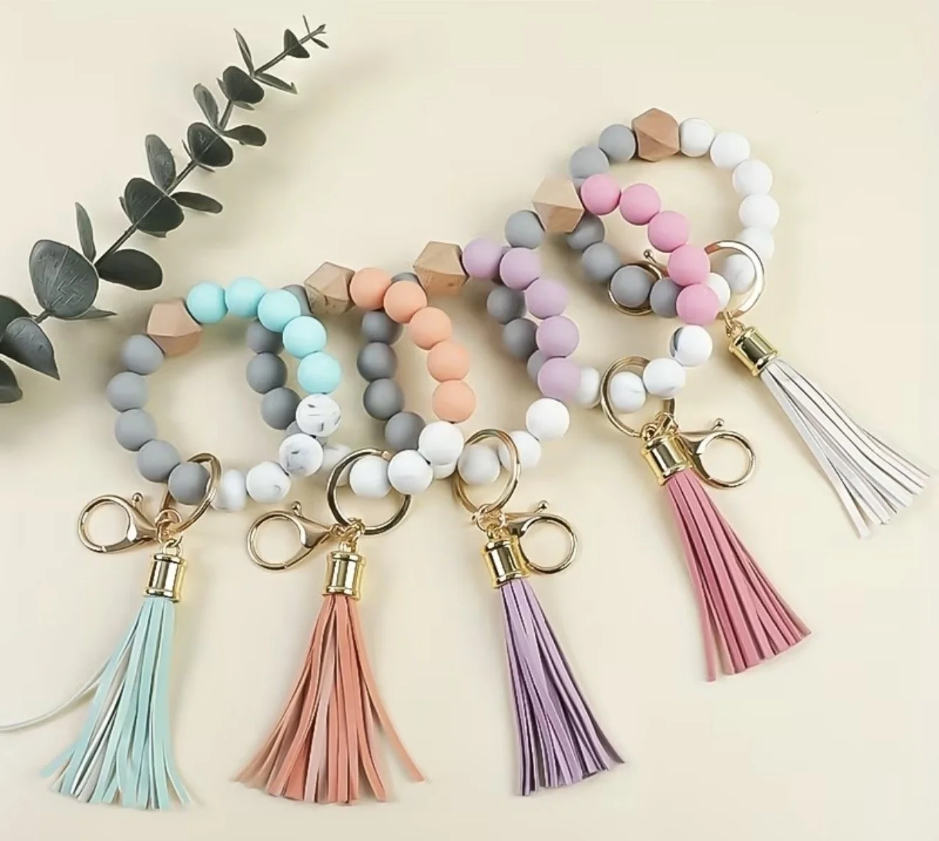 Colorful beaded keychains with tassels in pastel colors, arranged in a row on a light surface, with a green leafy branch on the side.