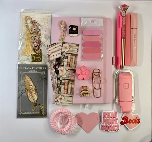 Pink and gold stationery and accessories, including sticky notes, paper clips, a keychain, a flower-shaped eraser, a brush pen, a pink jewelry piece, a pink speaker, and decorative stickers that say 'Read More Books' and 'Books', arranged on a white surface.
