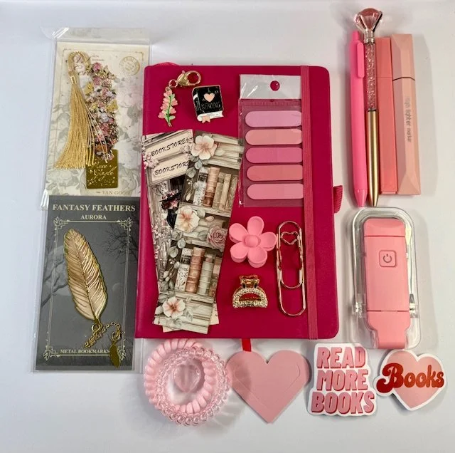 Flat lay of pink and gold stationery and accessories, including a pink notebook, pens, clips, stickers, a heart-shaped item, and decorative charms.