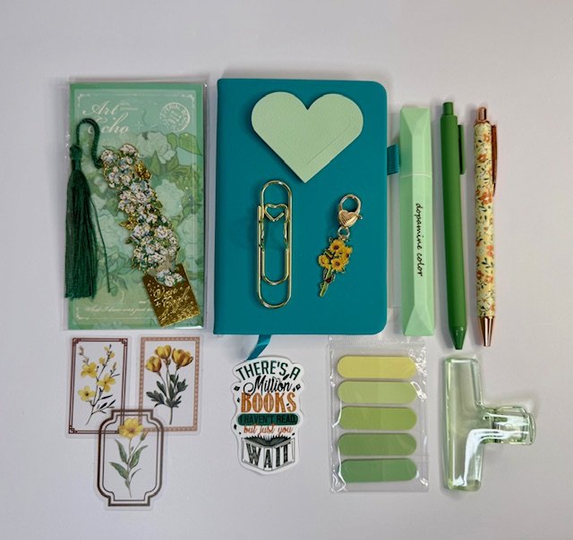 Flat lay of stationery items and decorative accessories, including a green bookmark with tassel, a blue notebook with a heart cutout, embellishments, pens, sticky notes, bookmarks, and decorative paper clips.