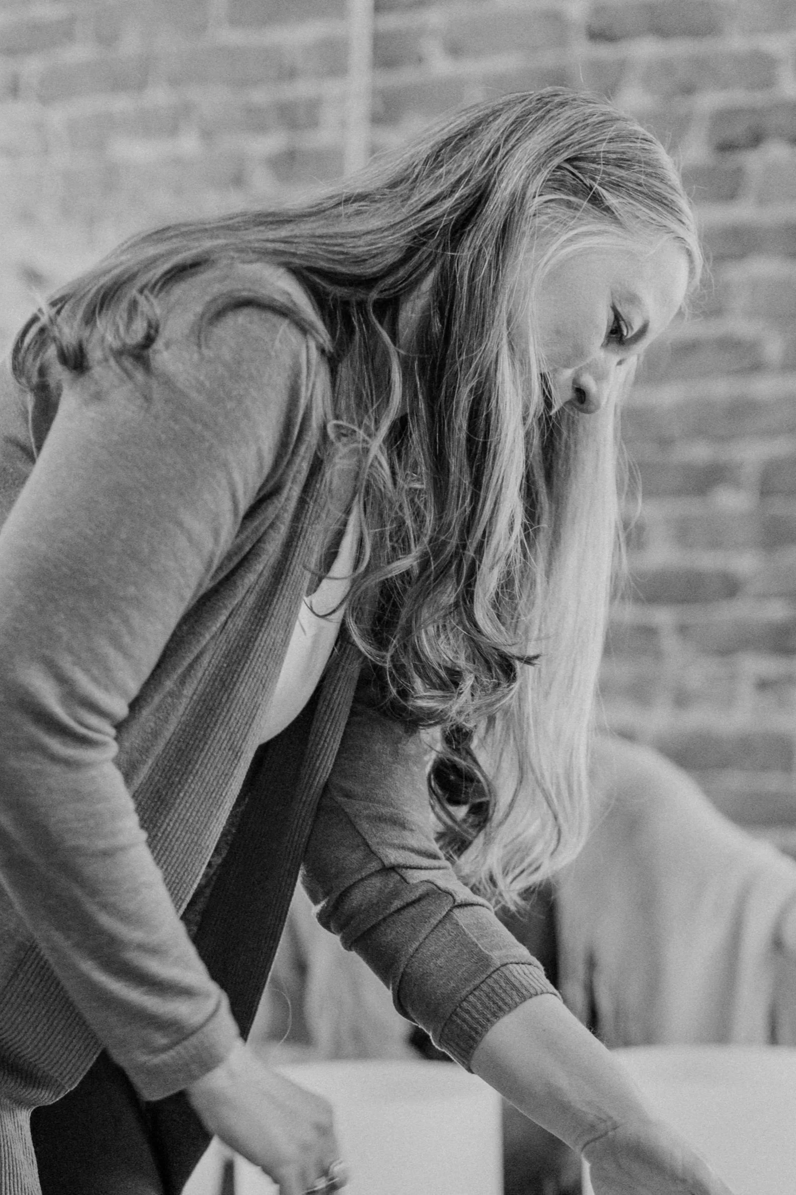 Woman with long hair bent over a table against a brick wall.