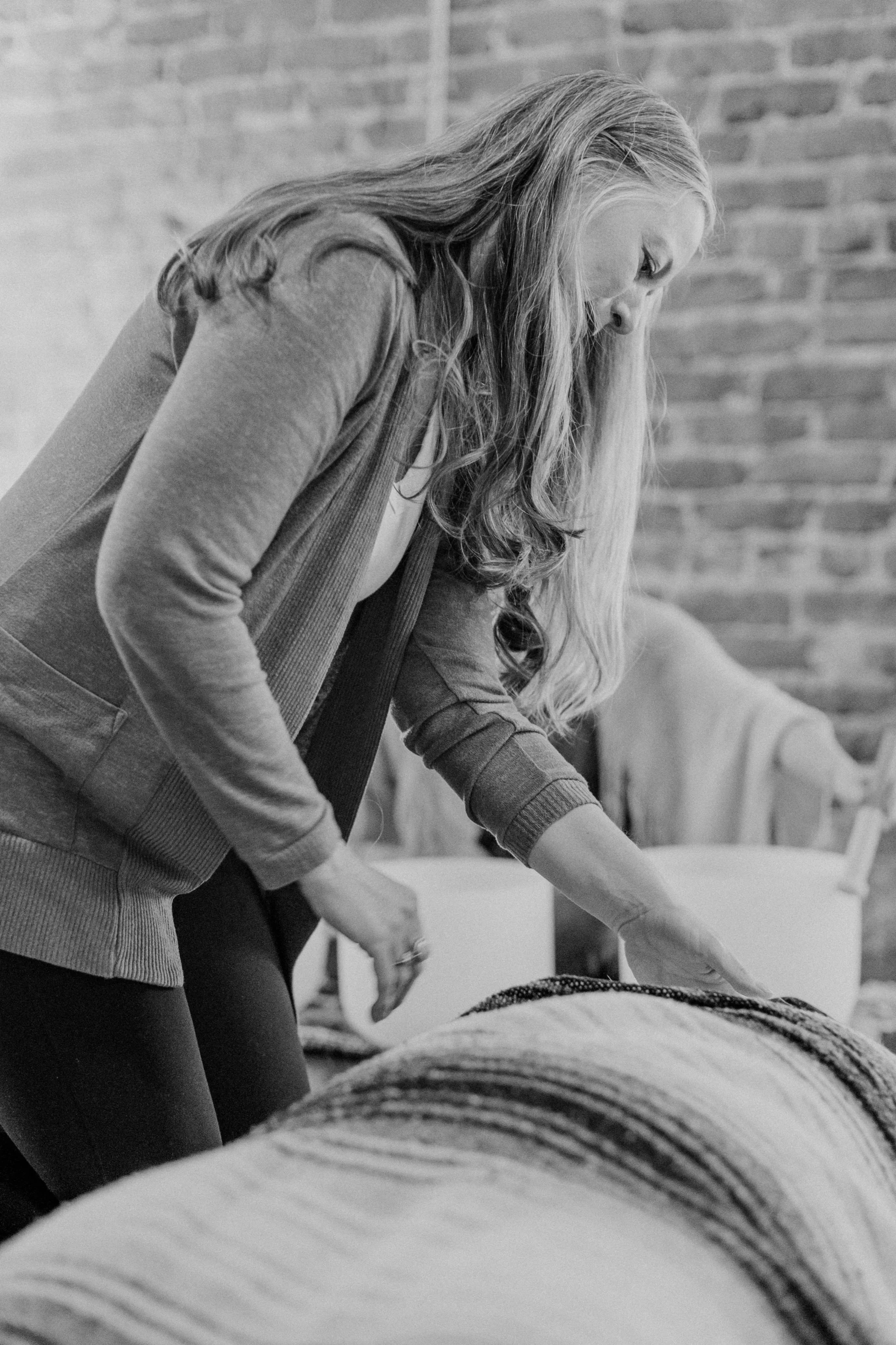 A woman with long wavy hair leaning over a bed, looking down at something. She is wearing a cardigan and dark pants, with a brick wall in the background.
