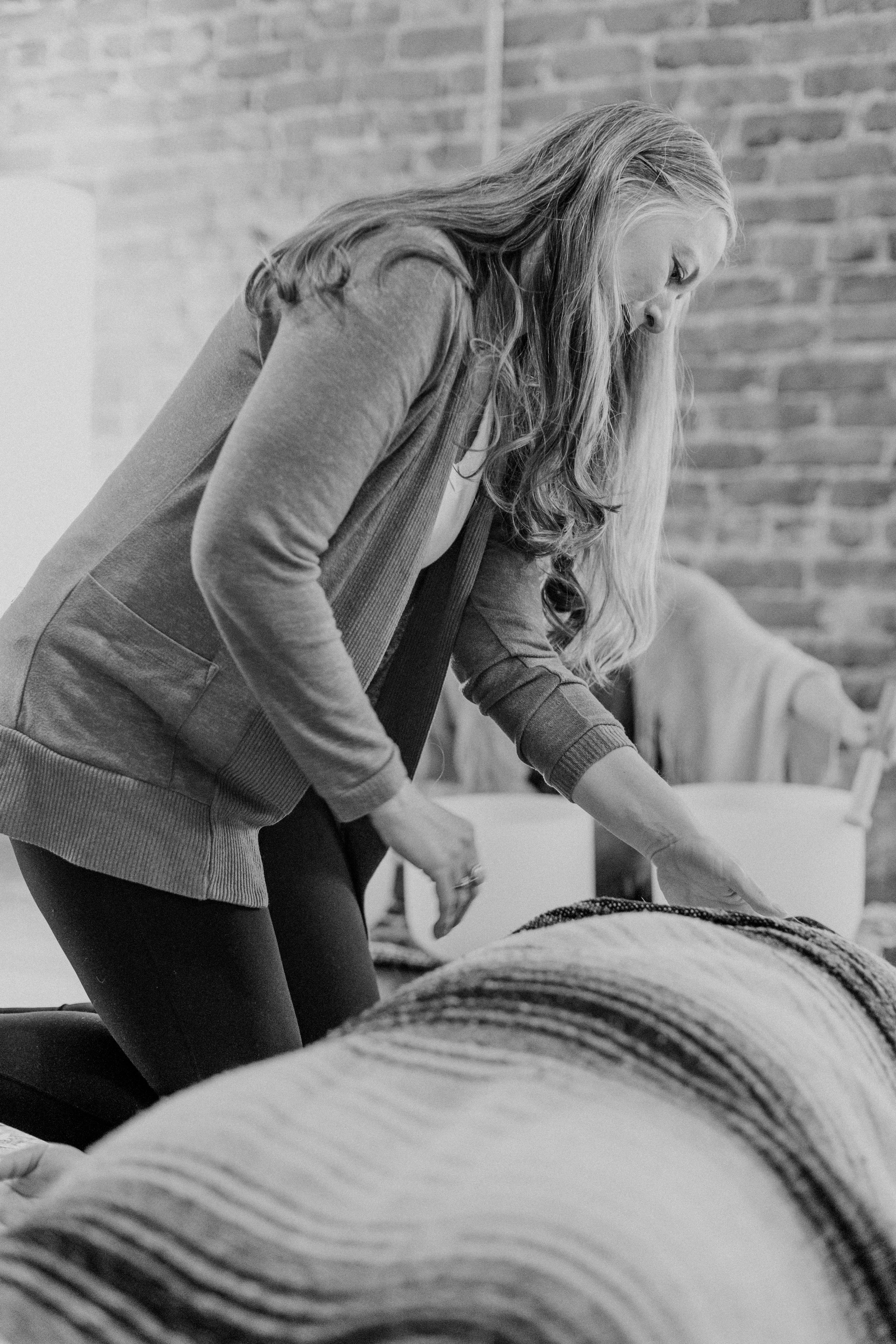 A woman with long hair leaning over a bed with striped blankets, in a room with a brick wall background.