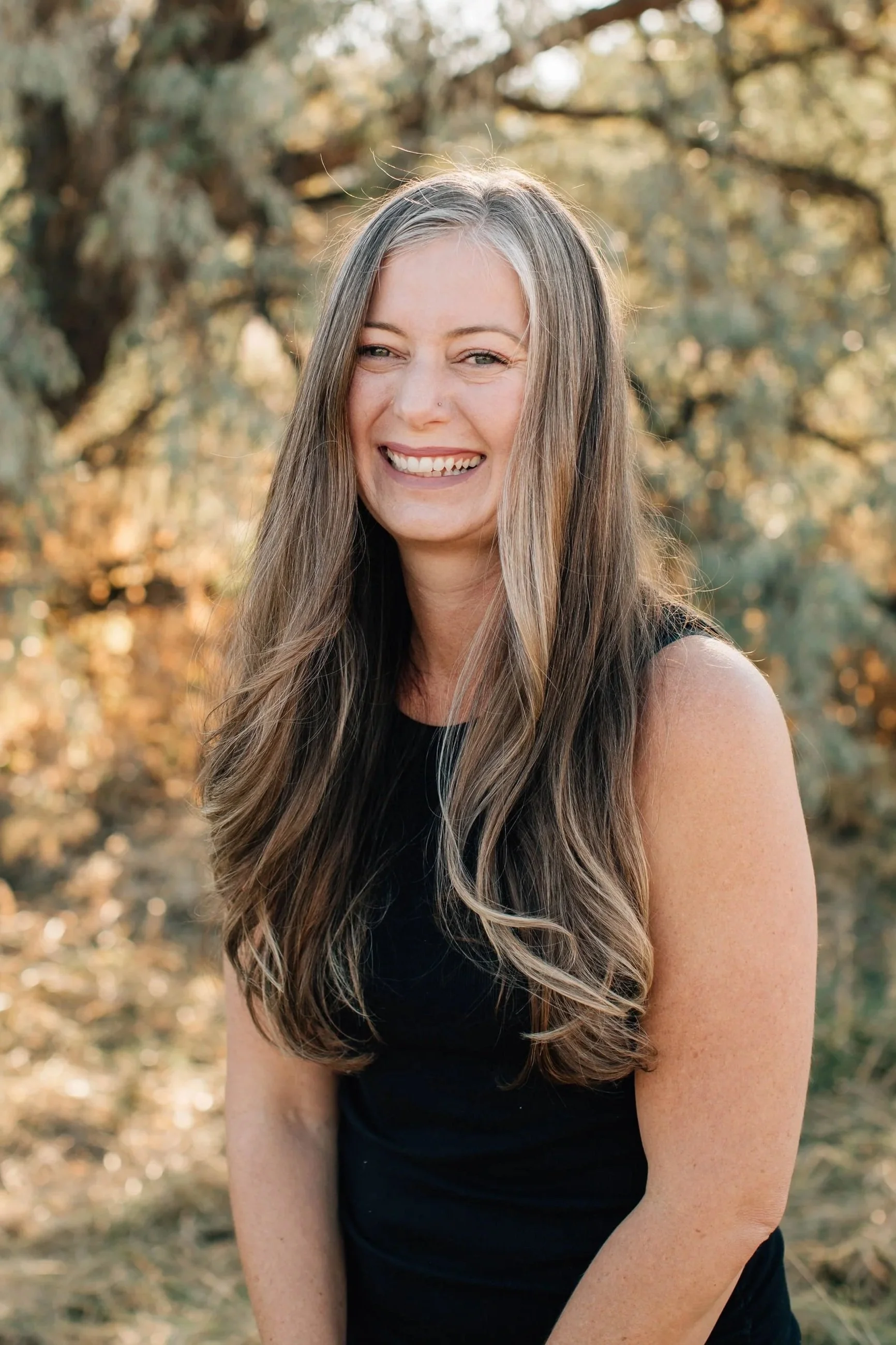 A woman with long, wavy hair smiling outdoors in a natural setting.