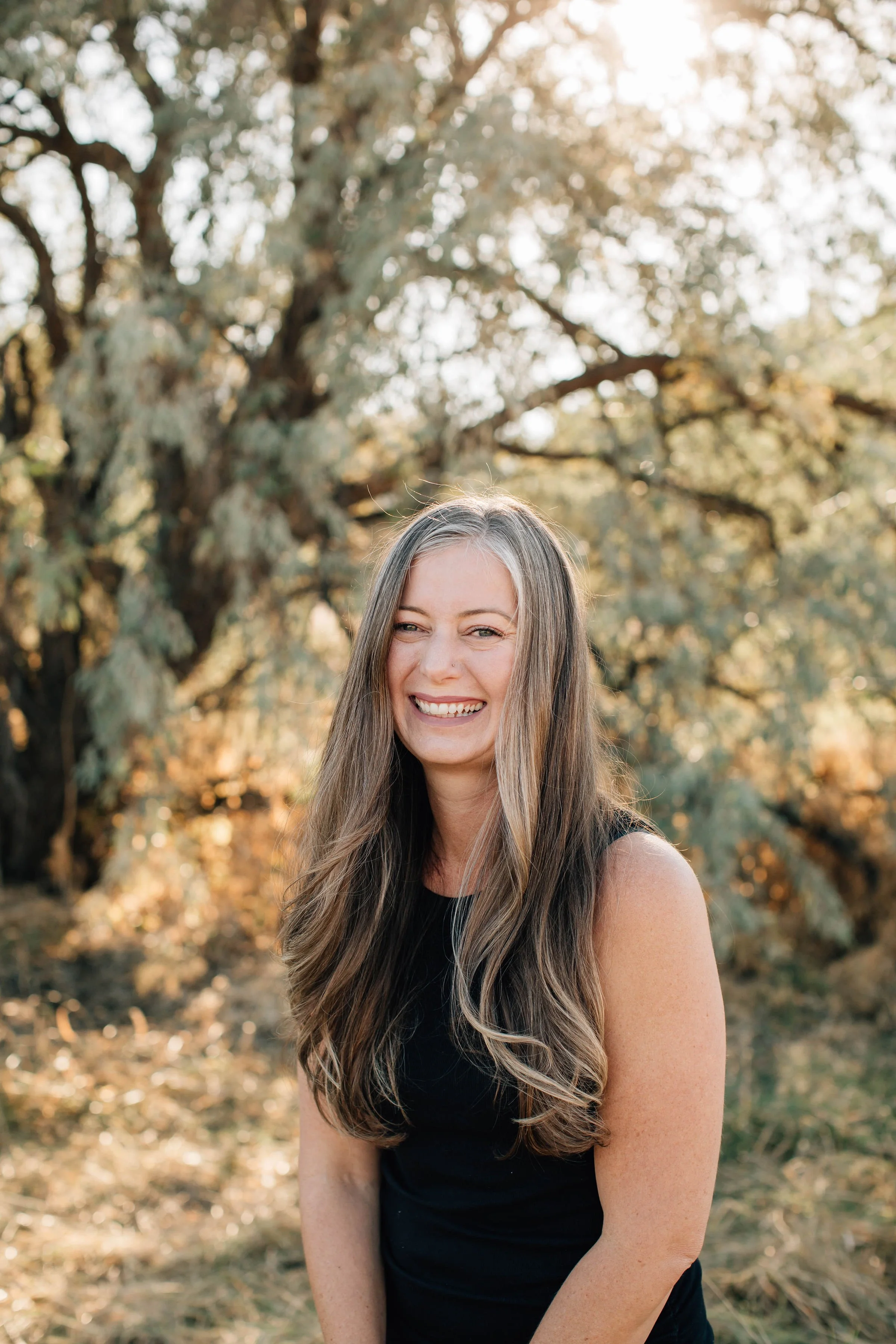 A woman with long, wavy hair smiling outdoors in front of a large leafy tree in warm sunlight.