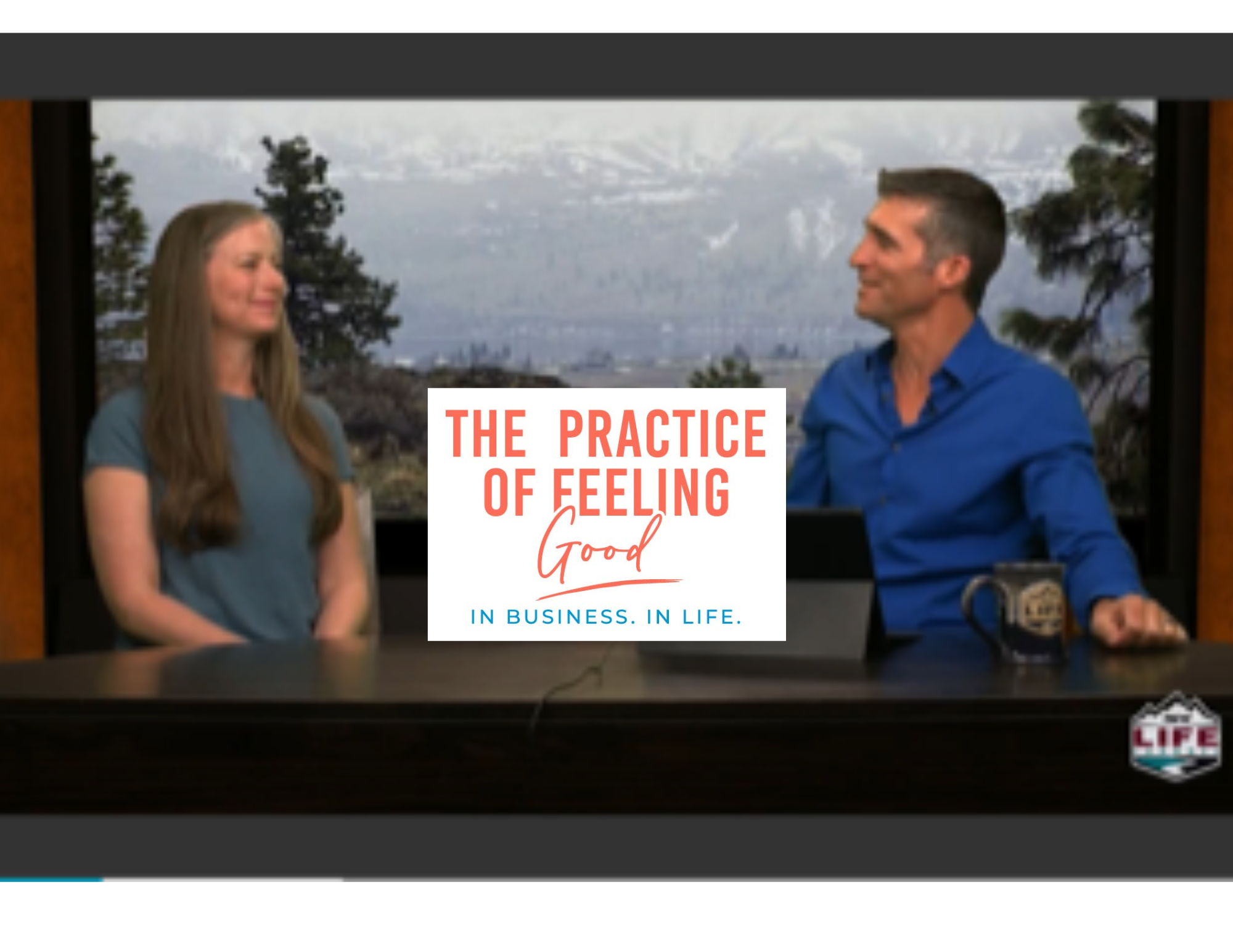 Two people seated at a table with a mountain landscape in the background. The person on the left has long, wavy hair and is wearing a short-sleeved blue shirt. The person on the right has short hair and is wearing a blue button-down shirt. There is a