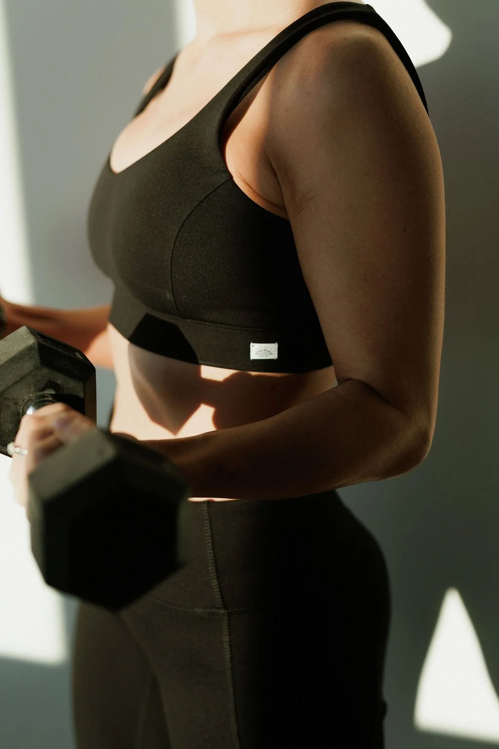 Close-up of a woman lifting dumbbells during workout, wearing a black sports bra and black leggings in a fitness setting.